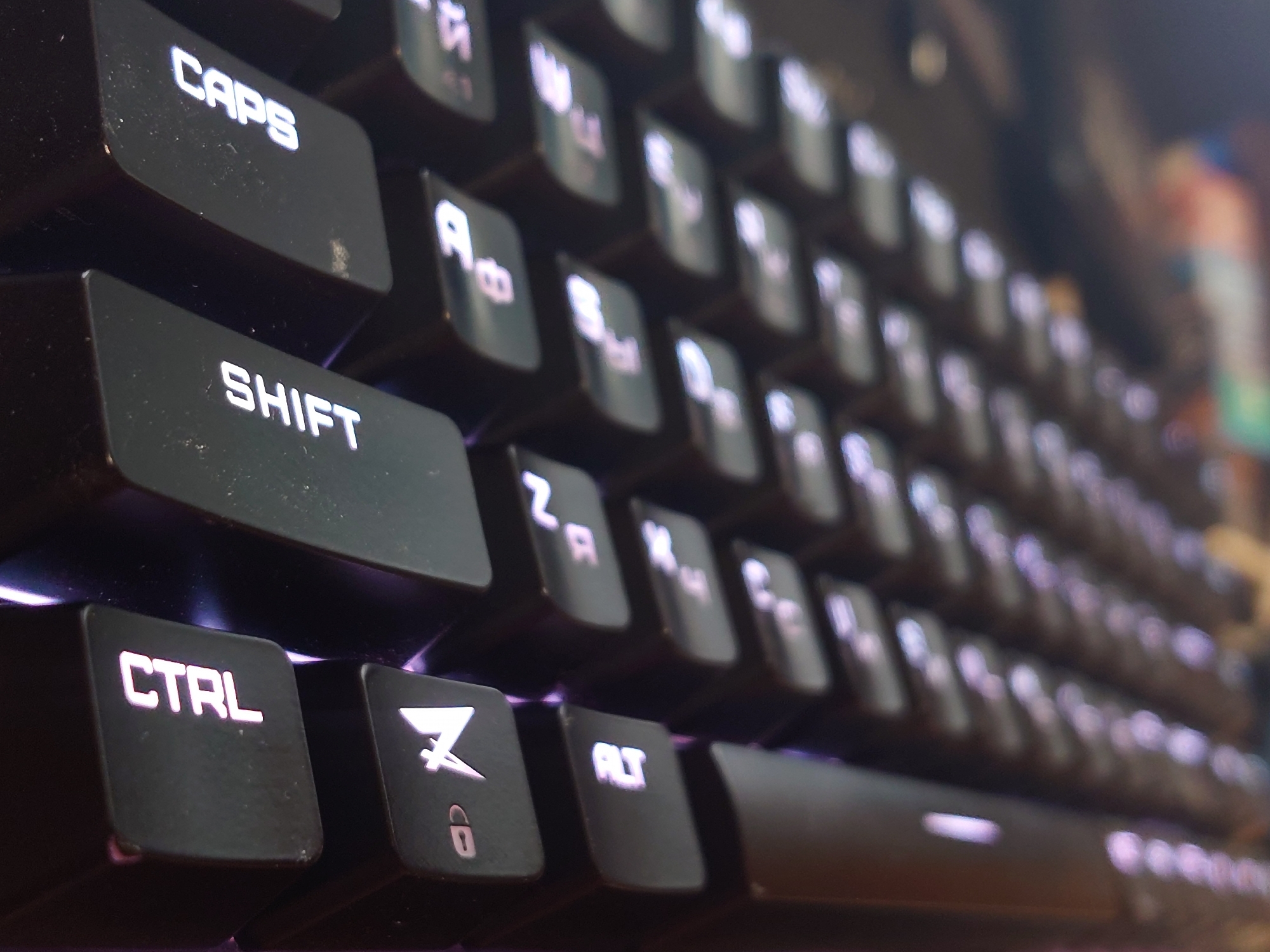 The image is a close-up, angled shot of a black keyboard with illuminated keys. The lighting is dim, with purple and white light emanating from the keys, suggesting a gaming or custom keyboard setup. The keys visible include "CAPS", "SHIFT", "CTRL", "ALT", and several other alphanumeric and symbol keys. The texture of the keys appears to be matte plastic. The background is out of focus and appears to be a desk or table surface, with blurred colors suggesting other electronic devices or a dimly lit room. The image is a still photograph and does not depict any people or actions. There is no discernible information in the image that would indicate its location in Sokolohirsk, Ukraine.