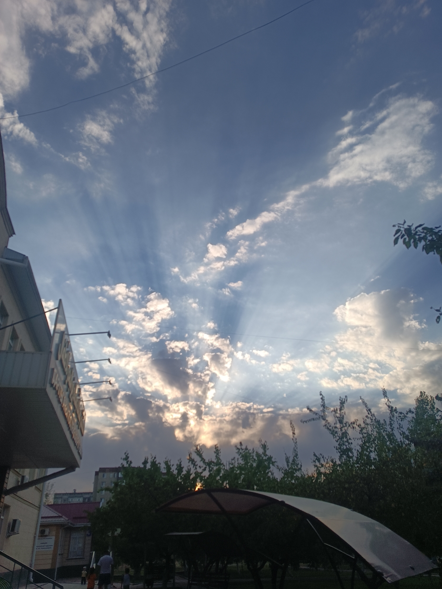 The image captures a dramatic sky with crepuscular rays piercing through clouds, indicating either sunrise or sunset. A portion of a building with signage is visible on the left, appearing to be a commercial establishment. In the foreground, a covered outdoor seating area with a translucent roof is present. Trees and other buildings are discernible in the background, suggesting an urban environment. The overall scene is set outdoors during daylight hours, with a clear indication of atmospheric light phenomena. The location is identified as Bishkek City, Kyrgyzstan.