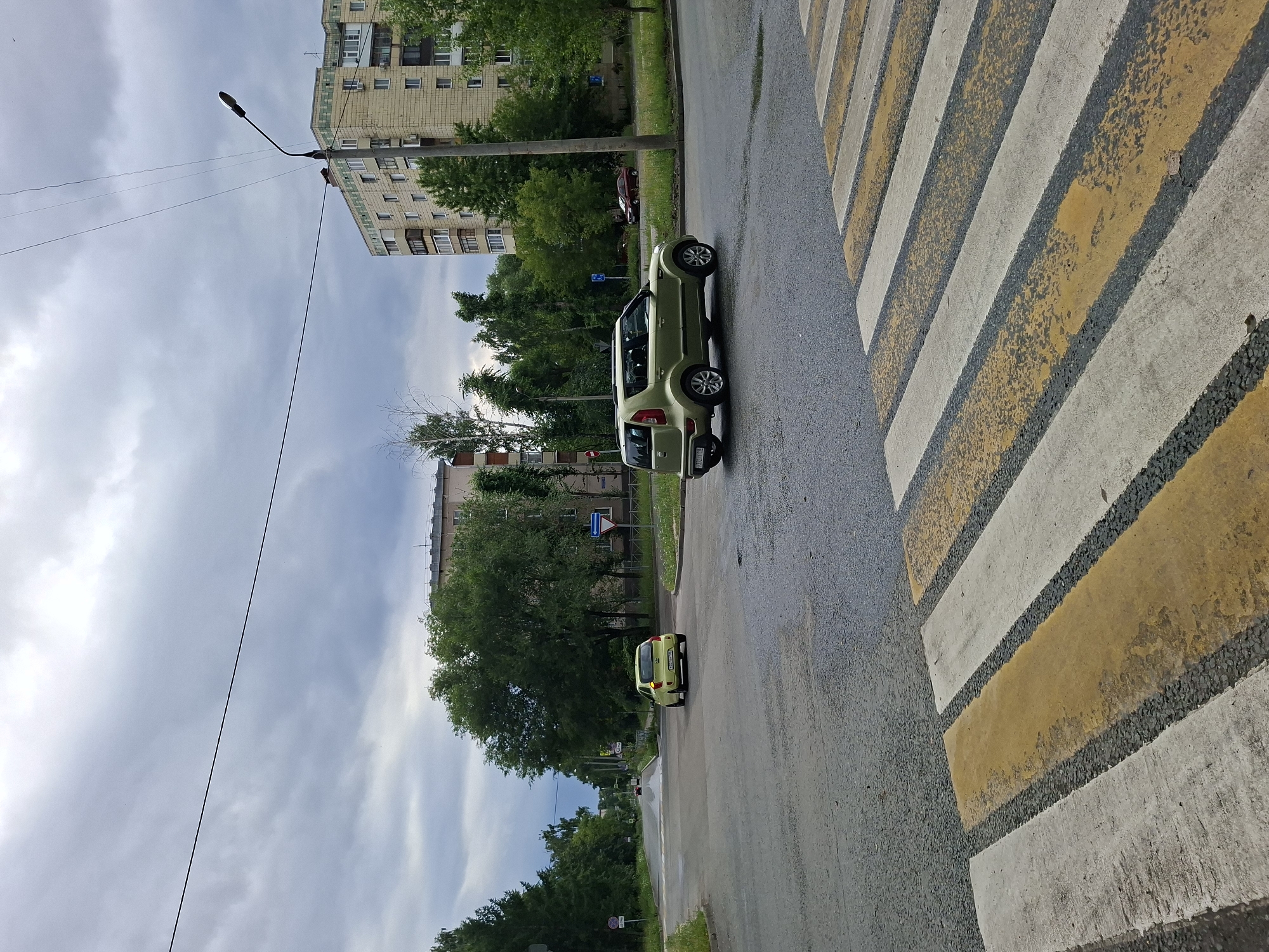 The image shows a street scene in Kazan, Russia, on an overcast day. A pedestrian crossing with yellow and white stripes is prominent in the foreground. Two light green cars are visible on the wet asphalt road. The first car, a compact SUV, is parked near the crosswalk. Further down the road, a smaller car is driving away from the viewer. Tall apartment buildings with many windows line the street, interspersed with lush green trees. A street lamp is visible in the sky. The clouds are heavy, suggesting recent rain or impending precipitation. The overall mood is calm and ordinary, depicting a typical urban environment. No people are visible in the image.