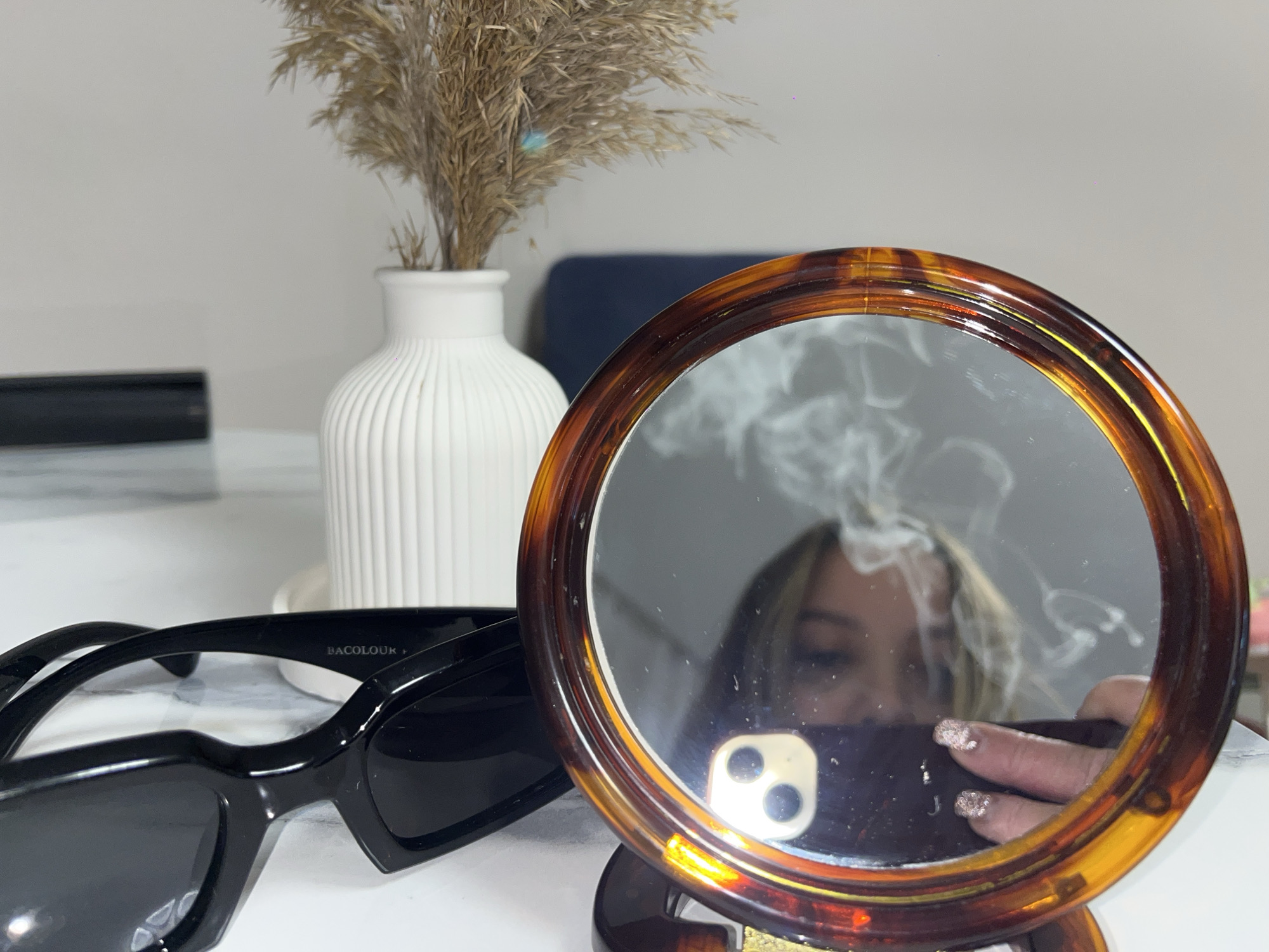 A close-up indoor shot reveals a still life arrangement on a light-colored surface in Krasnodar, Russia. The central focus is a round mirror with a tortoiseshell-patterned frame, propped upright. Reflected in the mirror is the upper face of a person with blonde or light-colored hair, holding a dark-colored smartphone with a visible dual-lens camera. The person's hand, adorned with glittery nail polish, is also reflected, appearing to hold the phone to capture an image. Swirls of white smoke or vapor are prominently featured in the reflection, emanating around the person's face.
To the left of the mirror, a pair of black, rectangular-framed sunglasses rests on the surface. The brand name "BACOLOUR +" is visible in white text on the inside arm of the sunglasses. In the background, a white, vertically ribbed ceramic vase holds a bouquet of dried, feathery pampas grass or similar botanical decor. A blurred dark blue object, possibly a piece of furniture, is visible further in the background. The lighting suggests an indoor setting, and no specific time of day or weather cues are apparent.