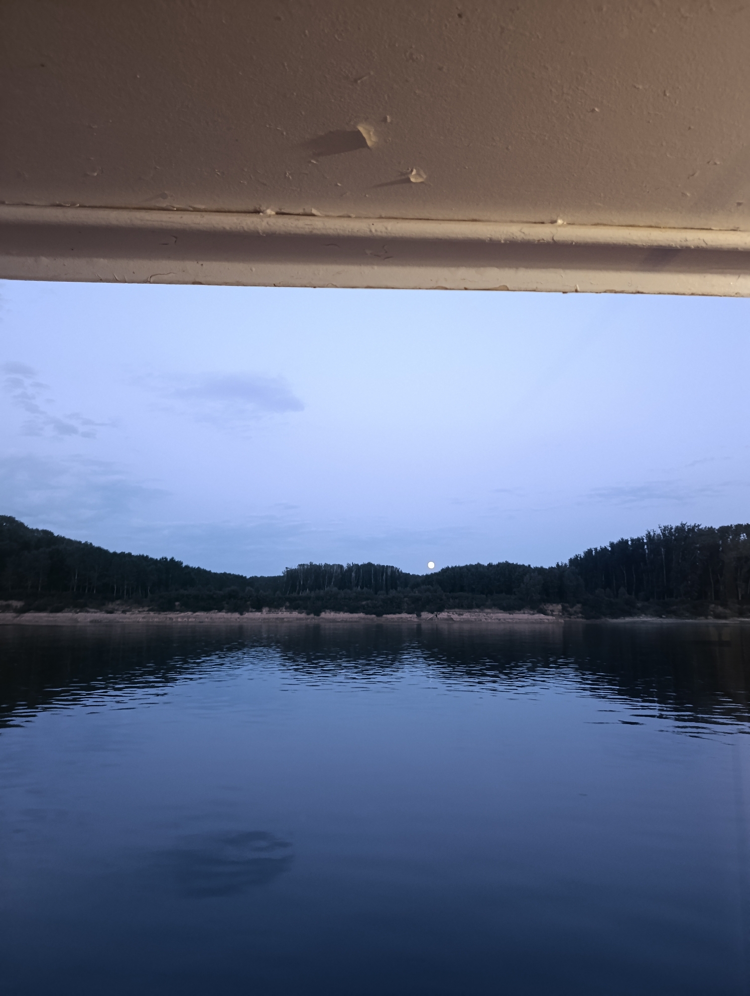 The image shows a tranquil view of a lake at dusk, framed by a light-colored overhang in the foreground. The sky is a soft, pale blue with a few wispy clouds and a small, luminous full moon visible in the distance. The water's surface is calm, reflecting the muted colors of the sky and the dark silhouette of a treeline that stretches across the horizon. The trees appear dense, forming a natural border to the lake. The overall impression is one of peacefulness and natural beauty. The specific location in the city of Slobozia, Romania, is not discernible from the visual information provided in the image.