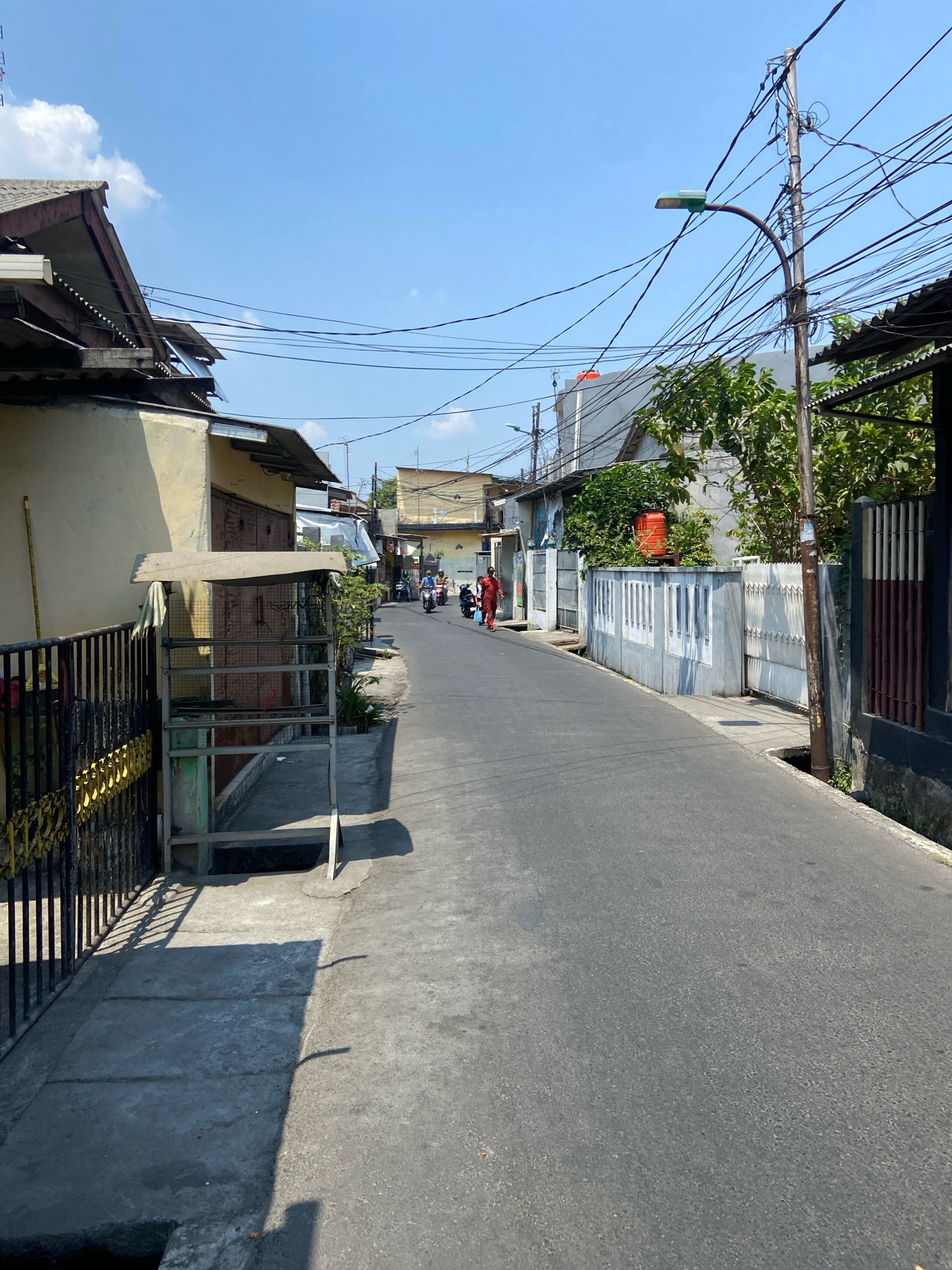 The image depicts a street scene in North Jakarta, Indonesia, under a clear blue sky. The street is paved and relatively narrow, with buildings lining both sides. On the left, a yellow-walled building with a corrugated metal roof is visible, featuring a black metal fence with yellow decorative elements. Adjacent to it is a freestanding metal structure with a sloped roof, possibly a vending cart or shelter, with a towel draped over it.
On the right side of the street, a white concrete fence with a repeating rectangular design separates the properties from the road. Behind the fence, lush green trees and an orange water tank are visible. A wooden utility pole with a dense network of electrical wires and a street lamp stands prominently.
Several people are present. Two individuals on motorcycles are moving down the street in the distance. Another person, wearing a red garment, is walking on the sidewalk on the left side of the road, heading away from the camera. The overall impression is that of a residential or mixed-use area in a tropical urban environment. The lighting suggests it is daytime, likely late morning or early afternoon, given the strong shadows cast by the buildings and objects.