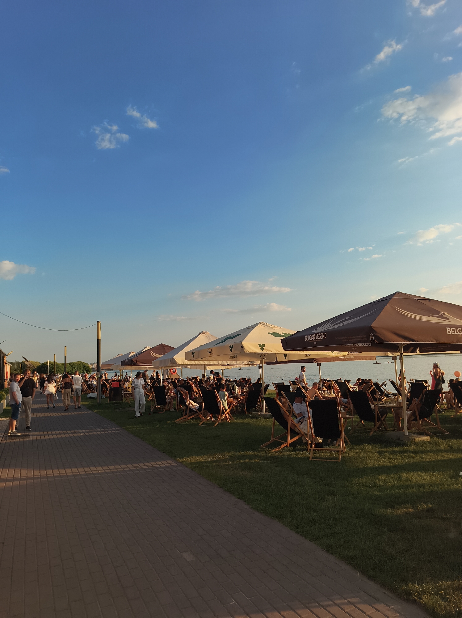 The image depicts a waterfront scene with a paved walkway on the left and a grassy area with seating and umbrellas on the right. The sky is blue with scattered white clouds. Several people are walking on the walkway and seated in lounge chairs under umbrellas. The umbrellas are predominantly white and dark brown, with one prominently displaying the text "BELGIAN LEGEND". The water body in the background appears calm, with distant figures and what might be paddleboarders. The lighting suggests it is late afternoon or early evening, with long shadows cast on the walkway. The location is identified as Ternopil, Ukraine.