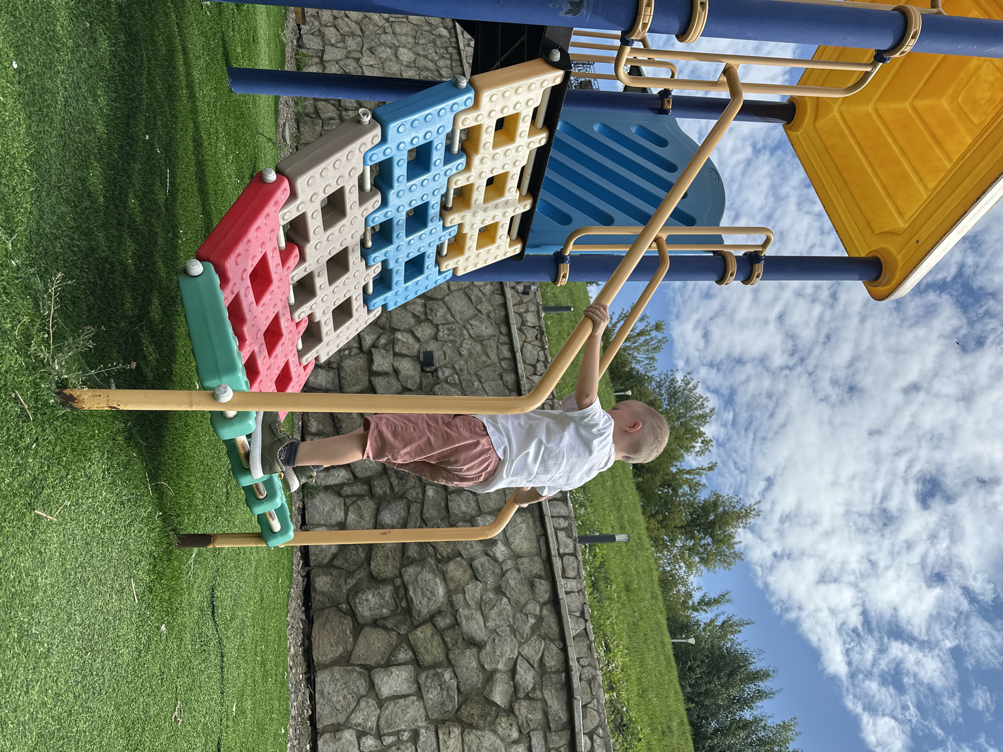 A young boy with blonde hair, wearing a white t-shirt and pink shorts, is climbing a playground structure. The structure features yellow and blue components with a yellow slide. The boy is holding onto a yellow railing. The playground is situated on bright green artificial turf, with a stone wall and a grassy slope in the background. The sky is filled with white clouds. The location is identified as Danilovka, Altai Krai, Russia.