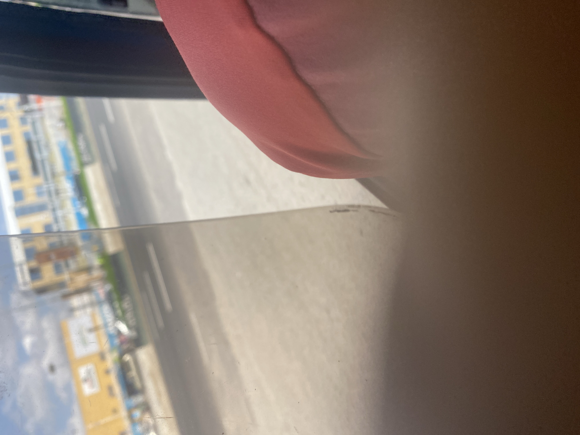 The image appears to be taken from inside a vehicle, possibly a car, looking out the side window. The window is slightly dirty. The outside scene is blurry, but appears to be an urban area with buildings, a road, and possibly some vehicles. There is a pink or reddish object partially visible in the upper part of the frame, possibly clothing or part of the vehicle's interior. The location is labeled as "Unknown, Unknown," indicating an unidentified city.