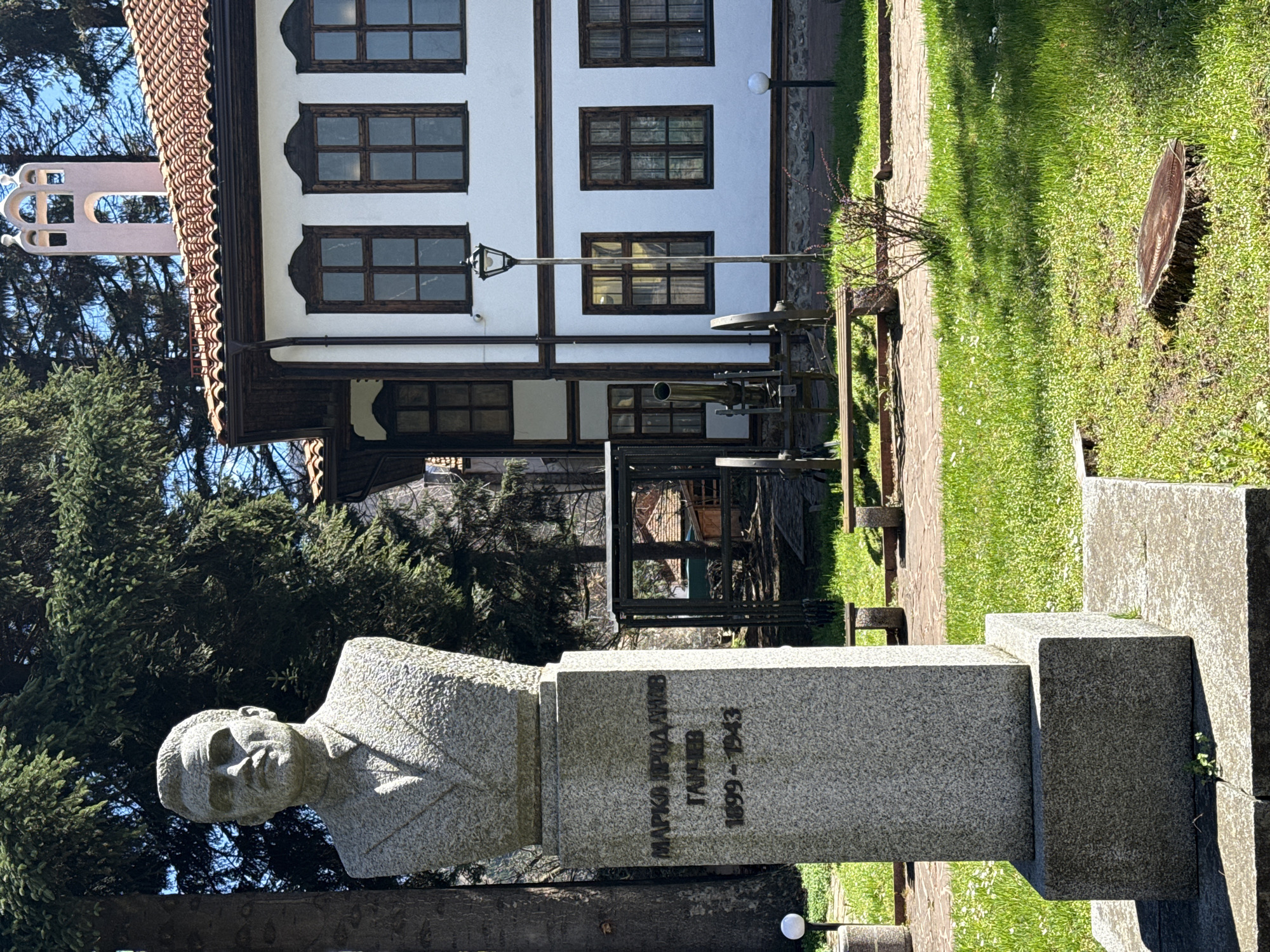 The image shows a monument of Marko Prodanić Galev, a Bulgarian revolutionary, who lived from 1899 to 1943. The monument is located in Etropole, Bulgaria. The monument consists of a stone bust of Galev on top of a stone pedestal. The inscription "MARKO PRODANIĆ GALEV 1899-1943" is engraved on the pedestal. In the background is a traditional Bulgarian house with white walls, dark wooden beams, and dark-framed windows. A street lamp and some metallic equipment, possibly associated with Etropole's history, are visible between the monument and the house. The surroundings appear to be a park with green grass and trees.