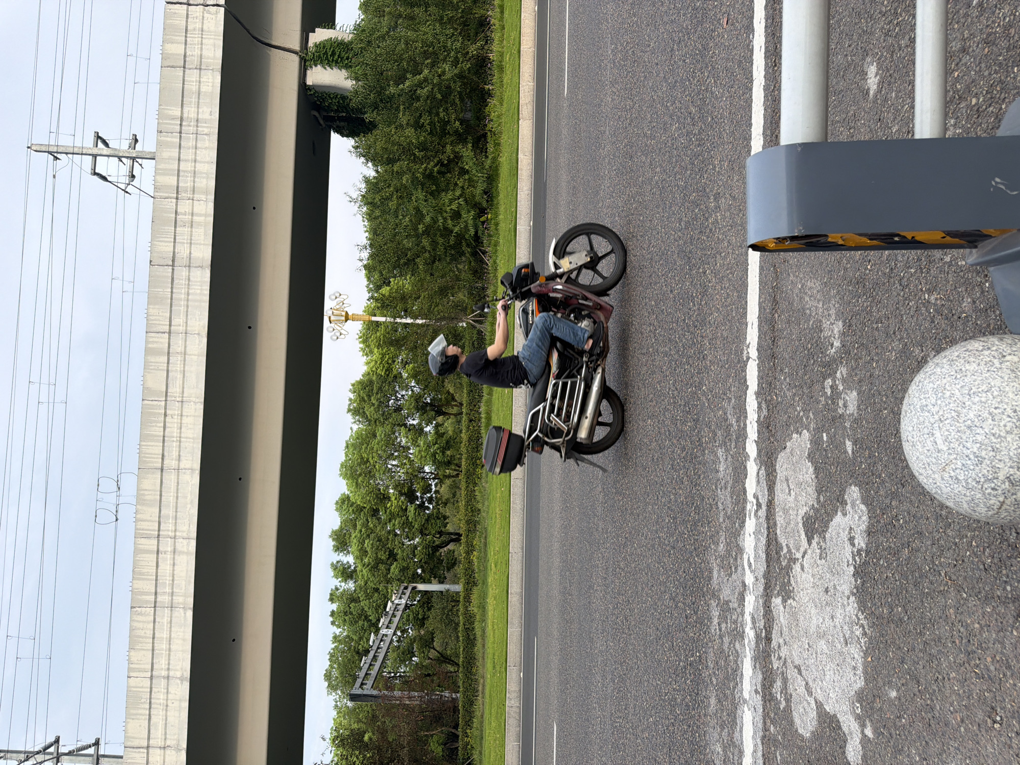 A person wearing a black t-shirt, blue jeans, and a helmet rides a motorcycle on an asphalt road under an elevated overpass in Jiangjin, China. The motorcycle is dark red and maroon with a black storage box on the back. Trees and greenery are visible behind a low hedge on the side of the road. Overhead, electrical wires are strung across poles, and part of the concrete overpass structure is visible, showing some staining and vines growing on a support pillar. The sky is overcast. In the foreground, a white dashed line is painted on the road, along with some weathered white markings. A grey, spherical bollard and a grey barrier with yellow reflective tape are also in the foreground.