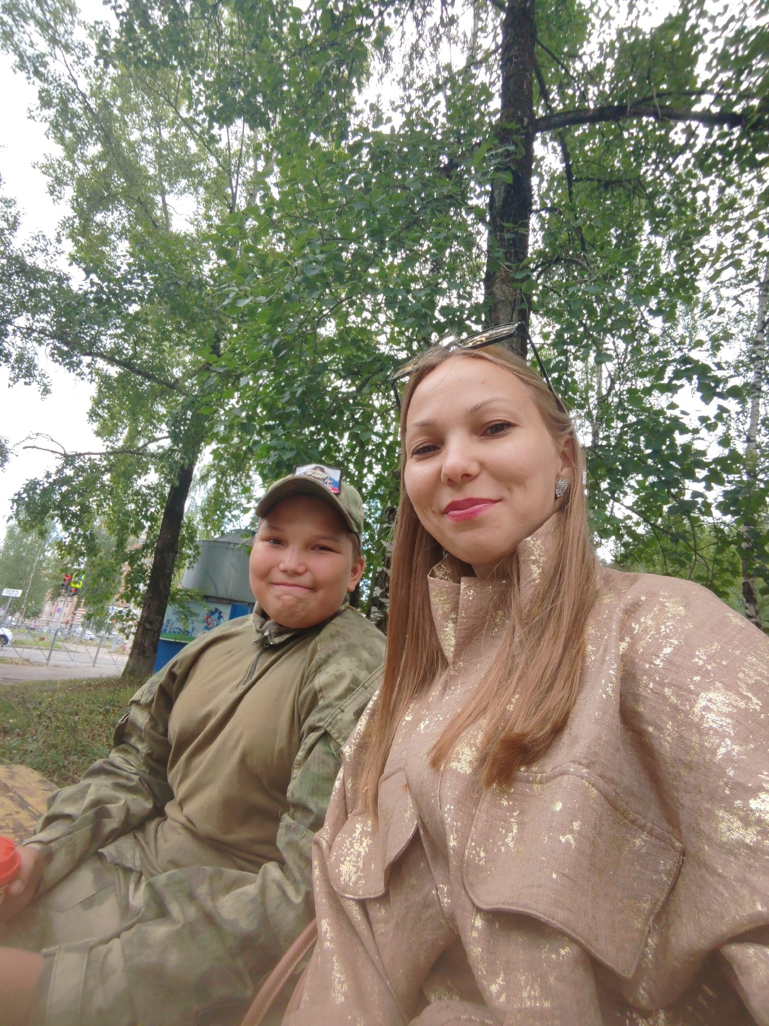 Two individuals are captured in a selfie-style outdoor photograph. On the left, a person wearing camouflage clothing and a baseball cap is seated. The cap has a patch on it depicting two figures. The person is smiling. To the right, a woman with long blonde hair, wearing a tan jacket with gold embellishments, is also visible. She is smiling and wearing earrings. They are positioned in front of trees and greenery. In the background, a street with a traffic light and buildings can be seen, indicating an urban setting. The overall scene suggests a casual outdoor gathering in the city of Syktyvkar, Russia.