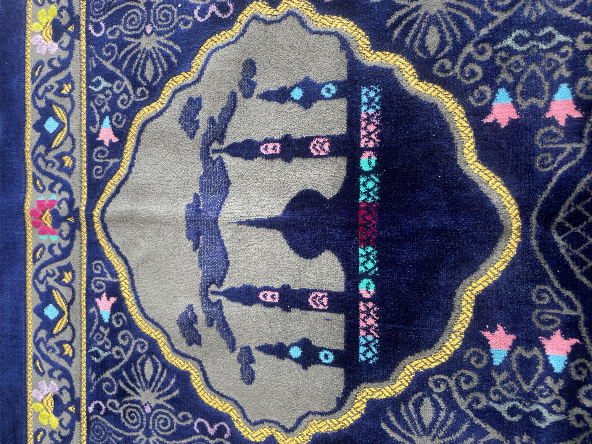 The image shows a close-up, overhead view of a navy blue prayer rug with intricate designs. The central motif features a silhouette of mosque minarets against a grey background, outlined by a wavy gold border. The minarets are decorated with pink and blue accents. The rug's outer edges and surrounding area are adorned with swirling grey patterns and small, colorful floral or star-like embellishments in pink, yellow, and blue. The texture of the rug is visible, appearing soft and possibly made of a plush material. The lighting suggests it is indoors. There is no visible indication of the location being Trieng Paloh, Indonesia, or any specific environmental context beyond the rug itself. The image is solely focused on the design and texture of the prayer rug.