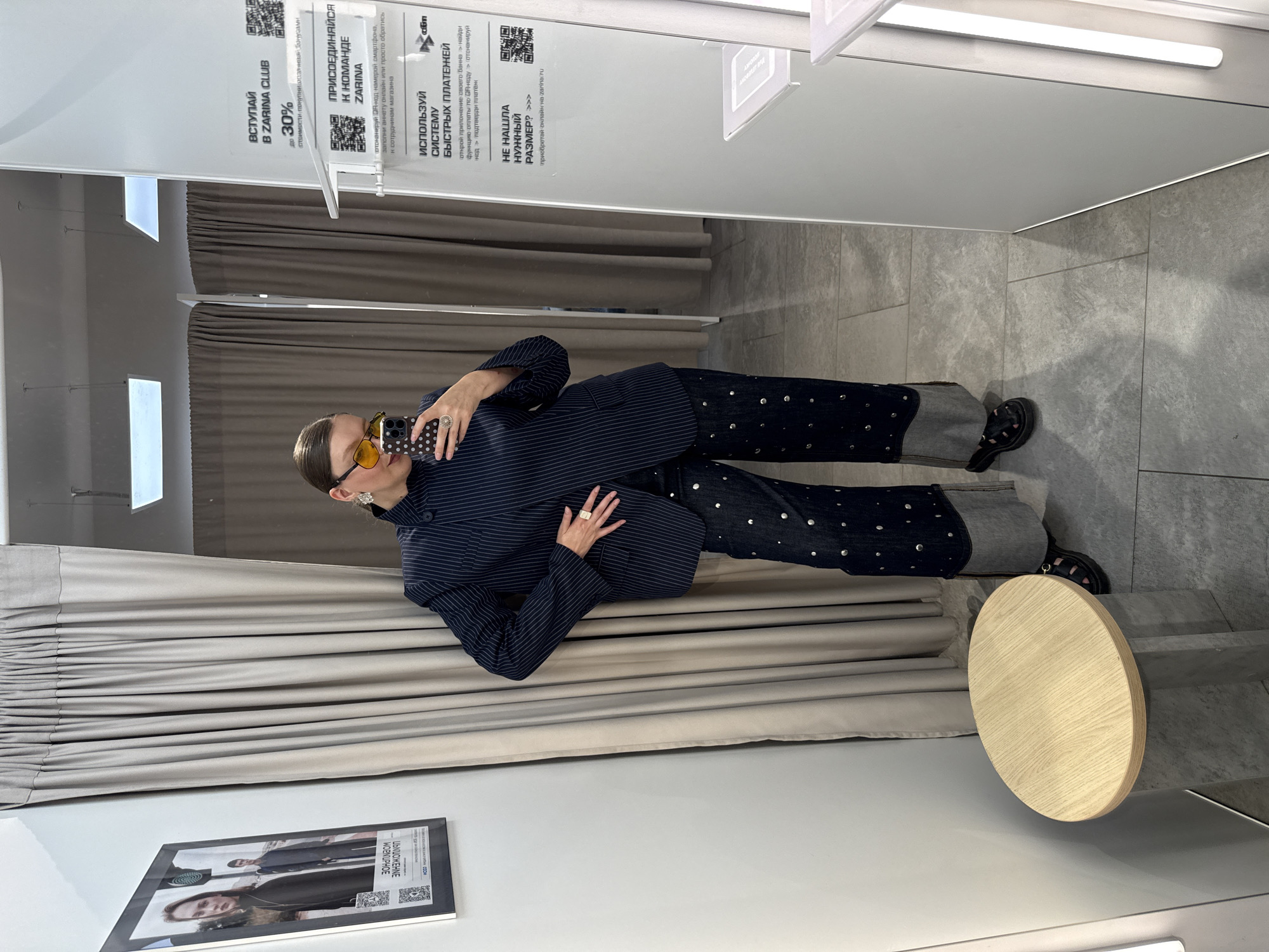 A woman is taking a selfie in a fitting room. She is wearing a navy pinstripe blazer, dark blue wide-leg jeans adorned with silver studs, and black sandals. She has on large, circular yellow-tinted sunglasses and a gold ring on her right index finger. Her hair is pulled back in a bun. The fitting room has light grey walls and light grey floor tiles. A beige curtain is visible to the left of the woman. A framed poster with two models is hanging on the wall to the left. A wooden stool with a grey base is to the woman's left. A full-length mirror reflects the woman and the fitting room. A sign on the mirror promotes the ZARINA CLUB and a quick payment system. The scene is indoors. Based on the information available, the fitting room is likely located in a ZARINA clothing store in Izhevsk, Russia.