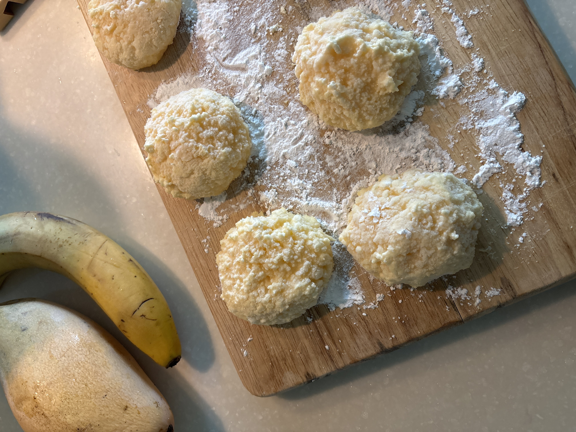 The image displays an overhead view of food preparation on a light wooden cutting board, likely in a kitchen setting in Choeng Thale, Thailand. Five unbaked, round food items, light yellow in color and with a textured, lumpy appearance, are arranged on the board. Each item is thoroughly dusted with a fine white powder, possibly flour or powdered sugar, which also scatters across the wooden surface.
To the bottom left of the cutting board, two fruits are visible on a light-colored countertop. The upper fruit is a ripe yellow banana, showing some natural blemishes and dark spots. Below it, partially obscured, is another elongated, light-colored fruit or vegetable, possibly a mango, given the tropical location context. The scene suggests a stage of food preparation, with the prepared dough balls ready for cooking or baking. No people are visible, and there is no discernible text in the image. The lighting is bright and evenly distributed.