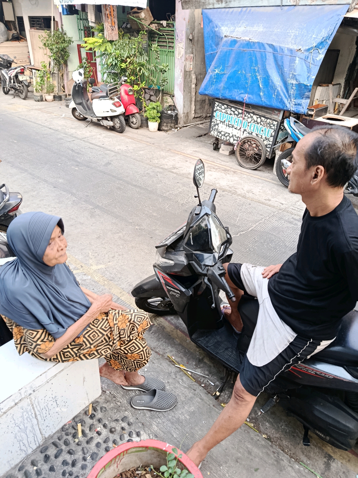 In North Jakarta, Indonesia, an elderly woman wearing a blue hijab and patterned skirt sits on a white ledge. To her right, a man in a black t-shirt and shorts sits on a black scooter, facing away from the camera. A second scooter, red and white, is parked behind them. In the background, a blue tarp covers a food cart labeled "Cappuccino Cincau." The street is paved and there are plants in pots. The scene depicts a casual street interaction in an urban environment.