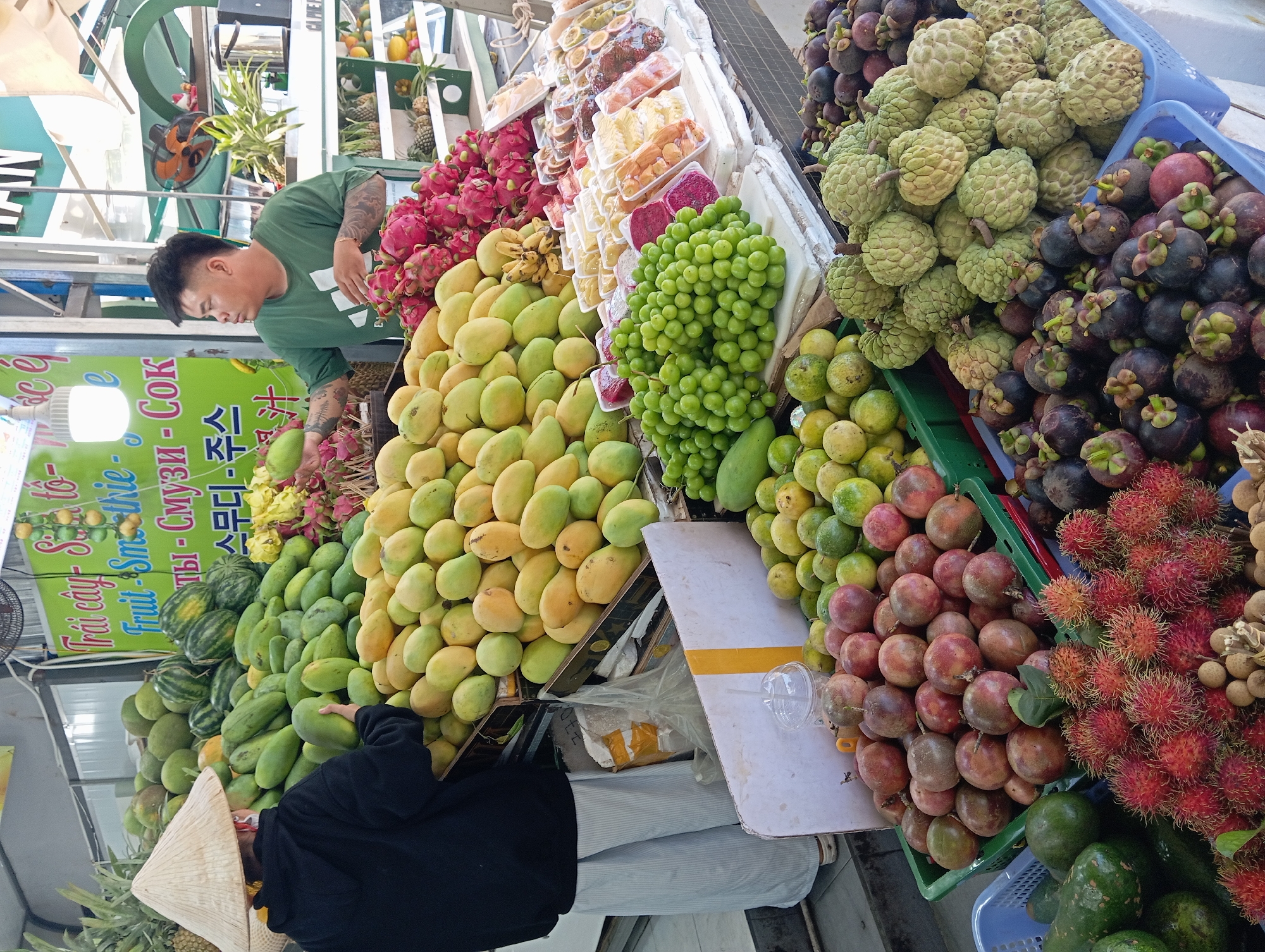 This is a street market stall in Nha Trang, Vietnam, displaying a large variety of fresh fruits. In the foreground, to the left, a person wearing a conical hat and a dark hoodie is standing in front of a display of green and yellow mangoes. To the right of the mangoes, a vendor in a green t-shirt is arranging fruit. The stall features numerous piles and baskets of fruits including dragon fruit, green grapes, rambutan, mangosteen, custard apples, passionfruit, oranges, and possibly watermelons and pineapples higher up. Packaged sliced fruits are also visible. The market appears to be outdoors or semi-covered, with various shelving units and signage visible in the background.
