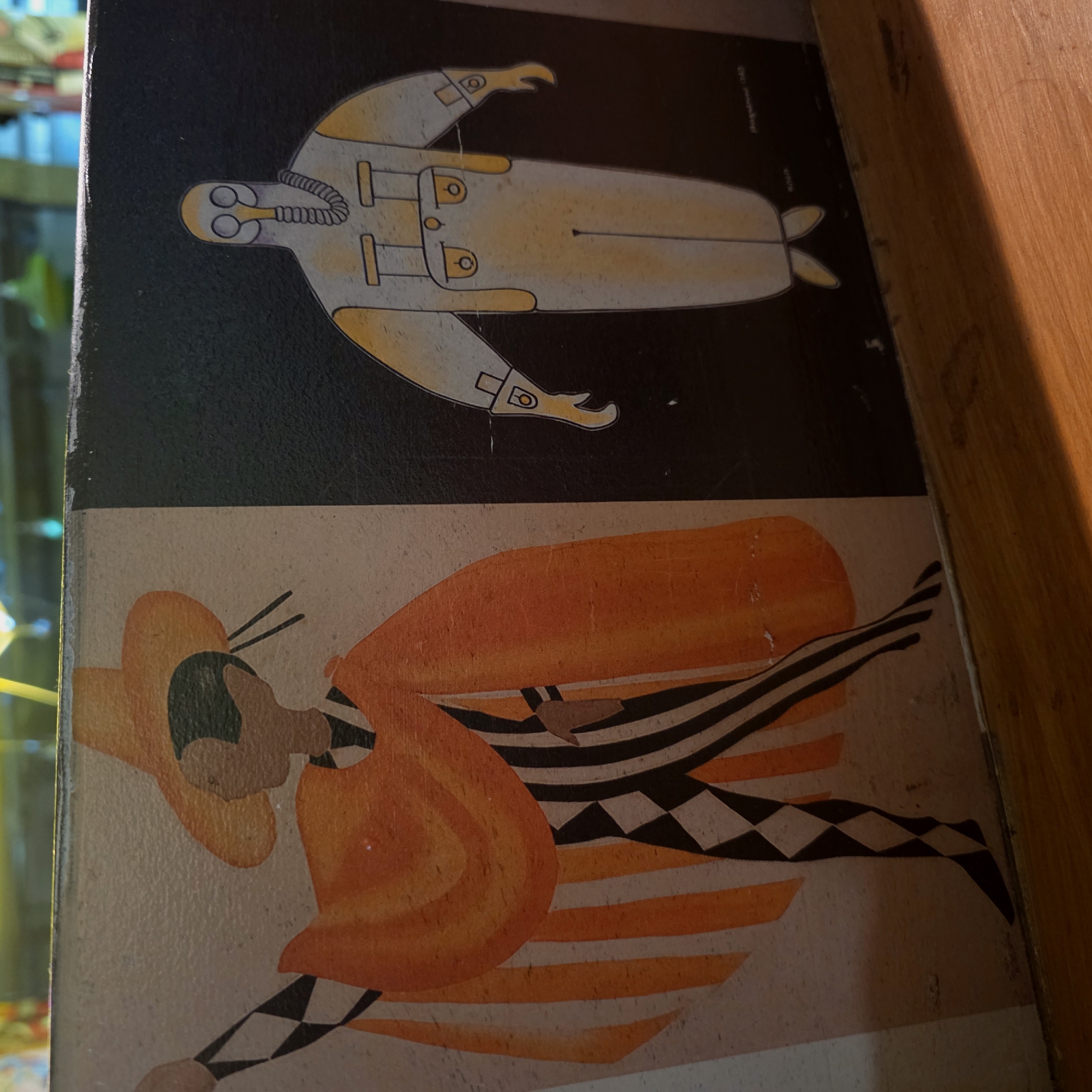 This image shows a vertical section of an object with two distinct illustrations. The top portion features a stylized drawing of what appears to be a deep-sea diver or astronaut in a yellow spacesuit against a dark background. The diver is depicted in a simplified, graphic style. Below, there's an illustration of a person wearing an orange outfit and a wide-brimmed hat. Black-and-white striped and checkered patterns are incorporated into the design, giving it an artistic, somewhat theatrical appearance. The exact location within Saint Petersburg, Russia, cannot be determined from the image.