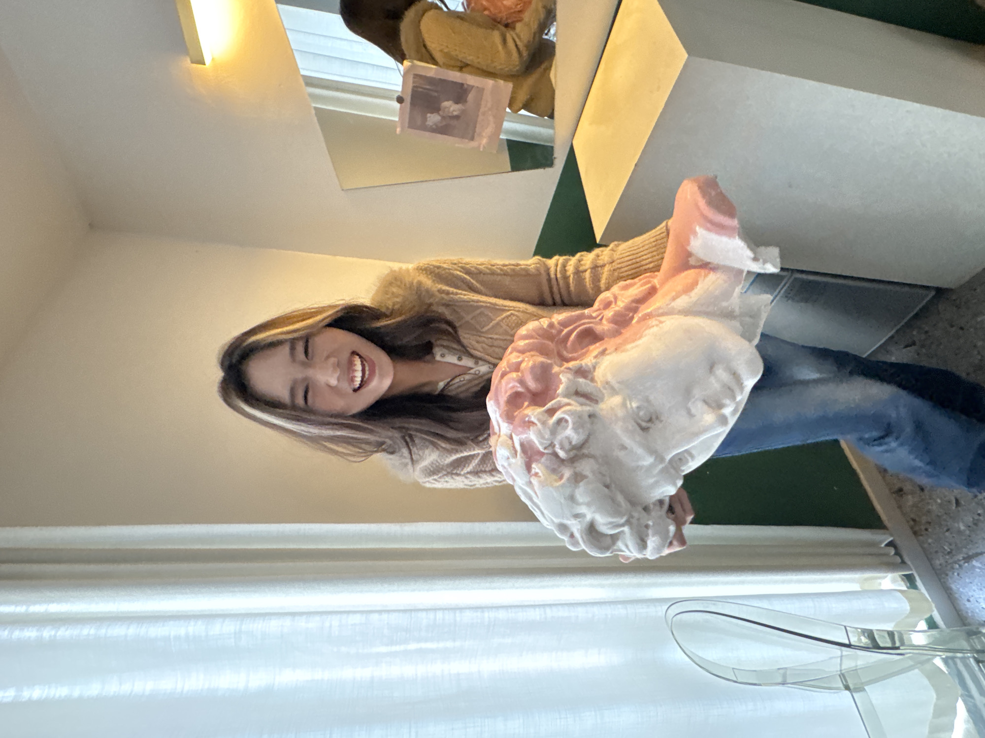 In the city of Liangjiang, China, a single adult female is depicted standing indoors, holding a large plaster bust. The female has long brown hair with blonde highlights and is smiling broadly with an open mouth. She is dressed in a beige ribbed cardigan, a light-colored collared shirt, and blue jeans. The object held is a white plaster bust of a classical male head with curly hair, which is notably colored pink on portions of the hair and upper face.
The surrounding environment features a room with light-colored walls in the upper sections and a dark green wall section at the lower right. On the left, sheer white curtains cover a window, admitting natural light. A transparent chair is partially visible near the curtains. On the right side of the image, a light-colored rectangular pedestal supports a wall-mounted mirror, to which a small, faded photograph is attached. A rectangular light fixture is mounted on the wall above the mirror. The floor has a speckled texture.