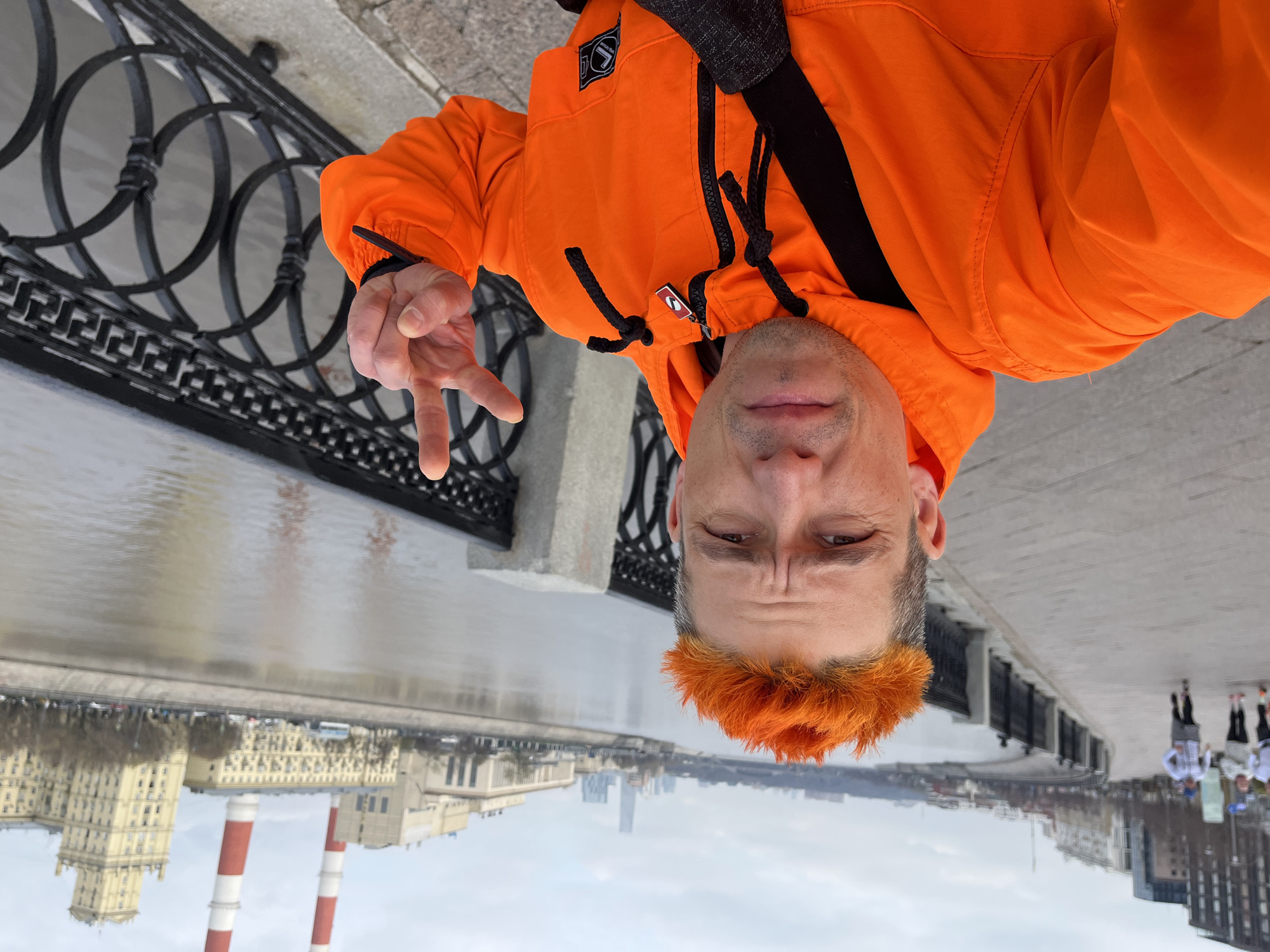 A man wearing an orange jacket and sporting orange hair is taking a selfie. He is standing on a bridge with an ornate black metal railing, holding up a peace sign. Behind him is the Moscow River, reflecting the city skyline and the cloudy sky. Visible in the background are industrial chimneys with red and white stripes. There's a building with classical architecture. A group of people in white and blue clothing can be seen further down the bridge. The man's head is positioned downwards in the frame. The photo appears to be taken in Moscow, Russia.