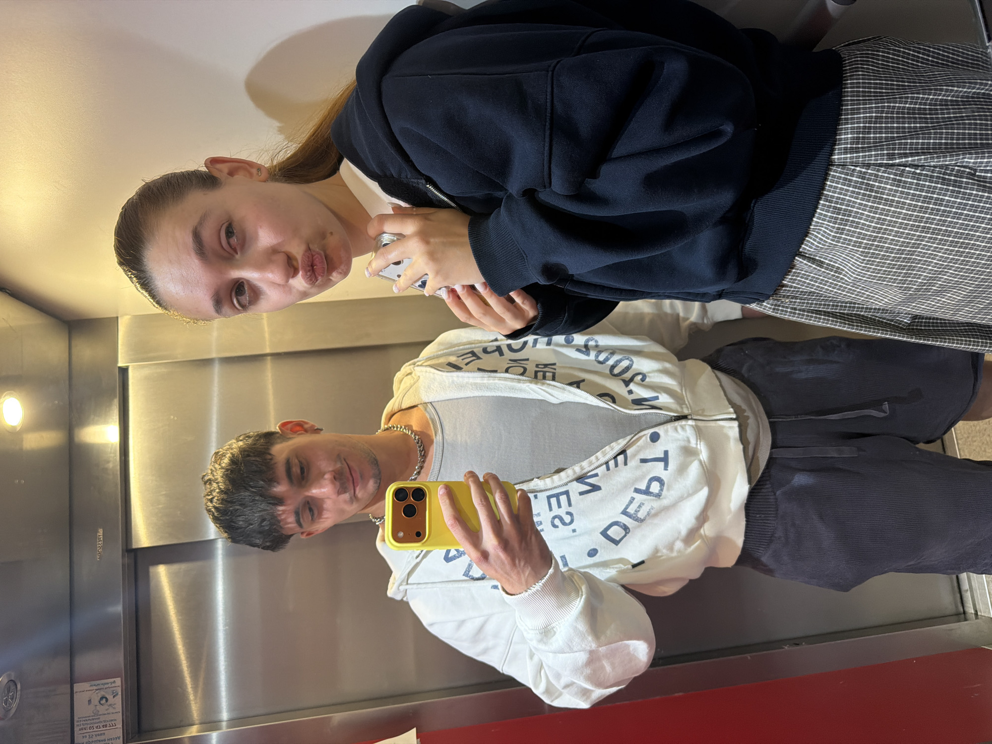 Two people are in an elevator taking a selfie. The man is holding a yellow phone and wearing a white jacket with text and gray pants. The woman is standing behind him, making a kissy face. She is wearing a navy hoodie and checkered pants. The elevator is metallic, and there is a red panel at the bottom. There is a sign in the lower left corner with Cyrillic writing. The scene is likely in Sofia, Bulgaria, due to the Cyrillic writing.