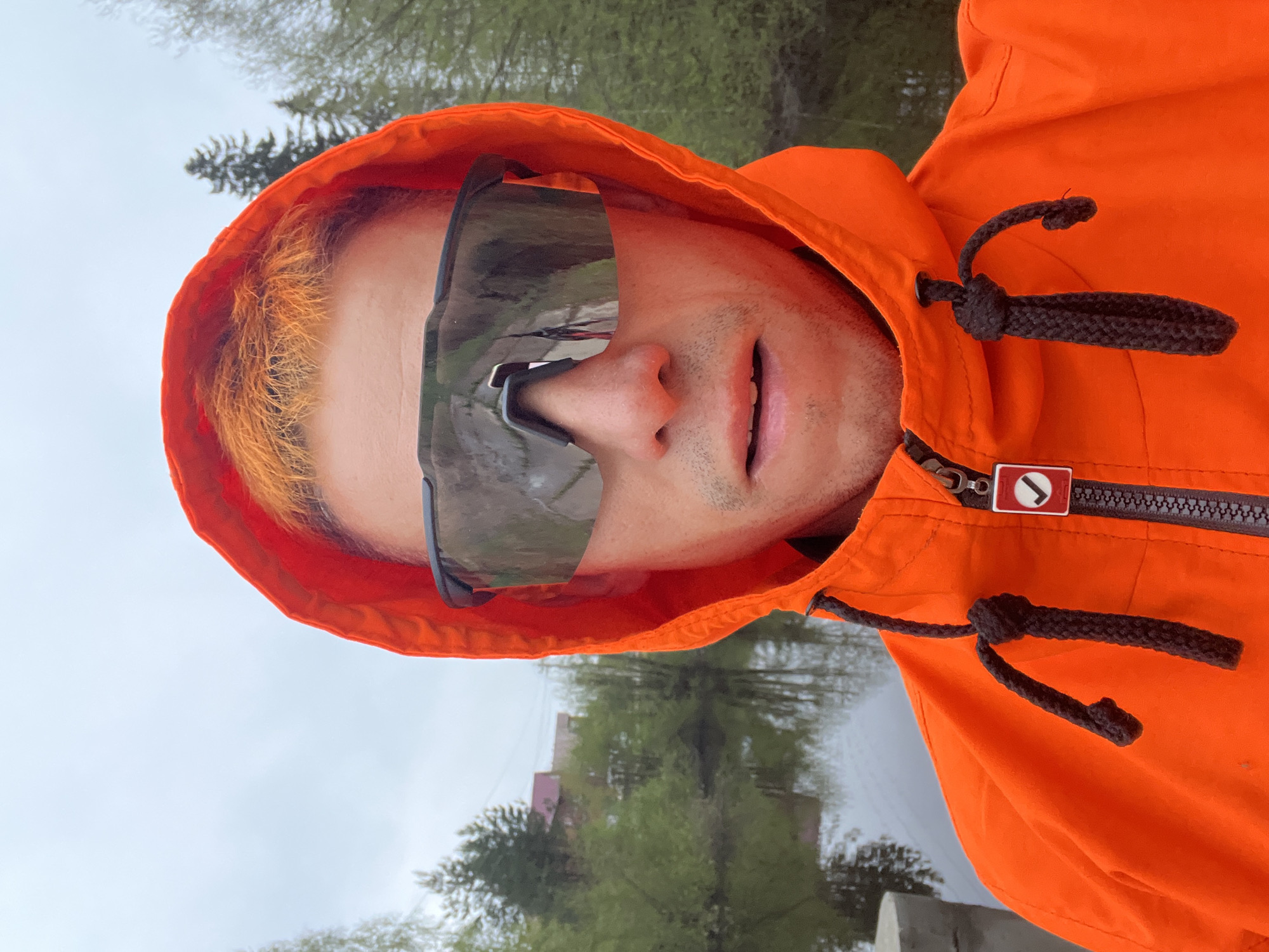 A person is wearing an orange hooded jacket and oversized sunglasses. The hood is up, revealing orange hair. The person is outdoors, near a body of water reflecting trees and a building. The location is Chaikovsky, Russia.