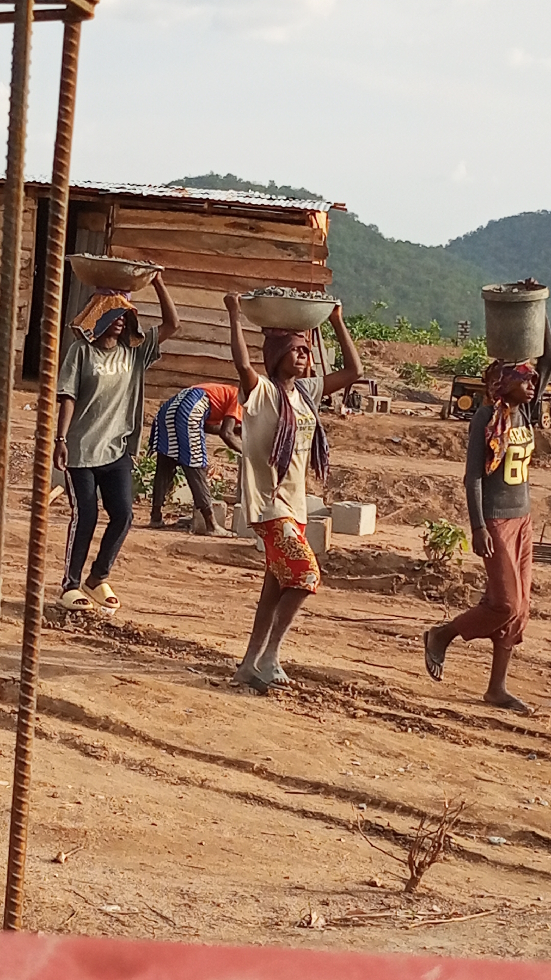 In Zango-ndaji, Nigeria, four individuals are engaged in construction-related labor at a site featuring a simple wooden structure and a portable generator. Three of the individuals are carrying bowls of material on their heads; two bowls appear to contain gravel, while the contents of the third bowl are obscured. The fourth individual is working on the ground near concrete blocks. The surrounding environment includes sparse vegetation and distant hills. The foreground shows construction materials and a dirt path.