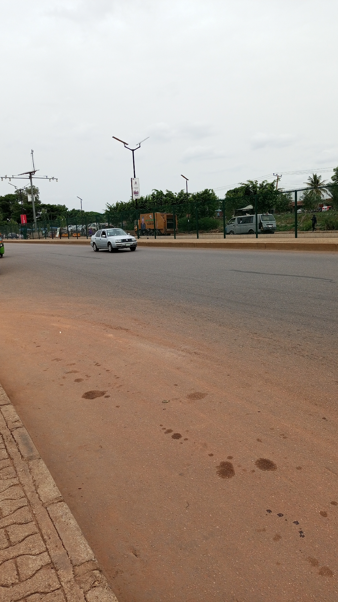 The image depicts a street scene in Kawo, Nigeria. A silver car is driving on a gray asphalt road with a yellow median. To the left is a red-brown pavement with several dark stains. A sidewalk with patterned stone is visible on the lower left. On the other side of the road, a fence separates the street from trees, a van, and a light-colored waste management truck. Utility poles with multiple lights and some signs stand near the background trees. A pedestrian is walking along the road. The sky is overcast.