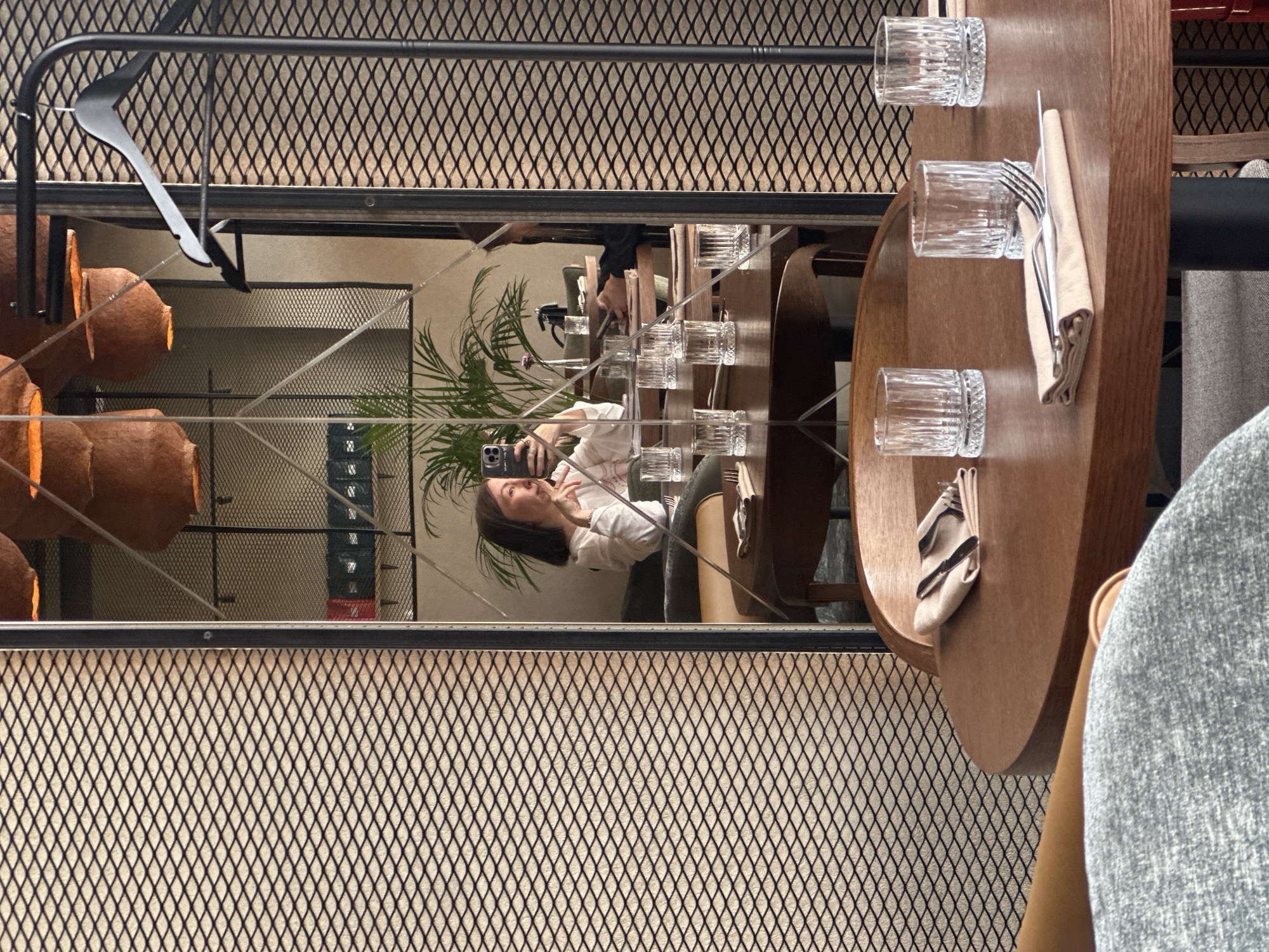 The image depicts the interior of a restaurant or cafe in Усады, Russia. A large, segmented mirror with a black frame occupies the central and right portions of the frame, reflecting various interior elements and a person. To the left and in the immediate foreground, a wall covered with a diamond-patterned metal mesh against a light background is visible.
In the foreground, a light grey upholstered chair partially obscures a round, polished wooden table. On this table, three empty, fluted glass tumblers are present. One tumbler is positioned adjacent to a folded beige linen napkin, which holds a silver fork and knife. A second similar napkin with cutlery is visible further back on the table.
The primary subject is an adult female, reflected in the mirror, taking a selfie with a smartphone. She has short, dark hair and is wearing a white long-sleeved top. The smartphone, held up to face level, exhibits a dual-camera module on its rear. Her mouth is slightly open. She is seated at a table set with multiple empty glasses, similar to those in the foreground. Behind her, a large potted plant with green fronds is prominently reflected.
The mirror's reflection additionally shows several other tables, some set with similar glassware, and chairs with dark upholstery and wooden backs. Overhead, multiple large, inverted, terracotta-colored conical light fixtures or decorative elements are suspended from the ceiling. A section of a wall with shelving holding dark, rectangular items is reflected in the background. On the far right of the reflection, a black metal coat stand with two black hangers is visible. The mirror's surface is intersected by thin, light-colored lines, indicating its segmented construction.