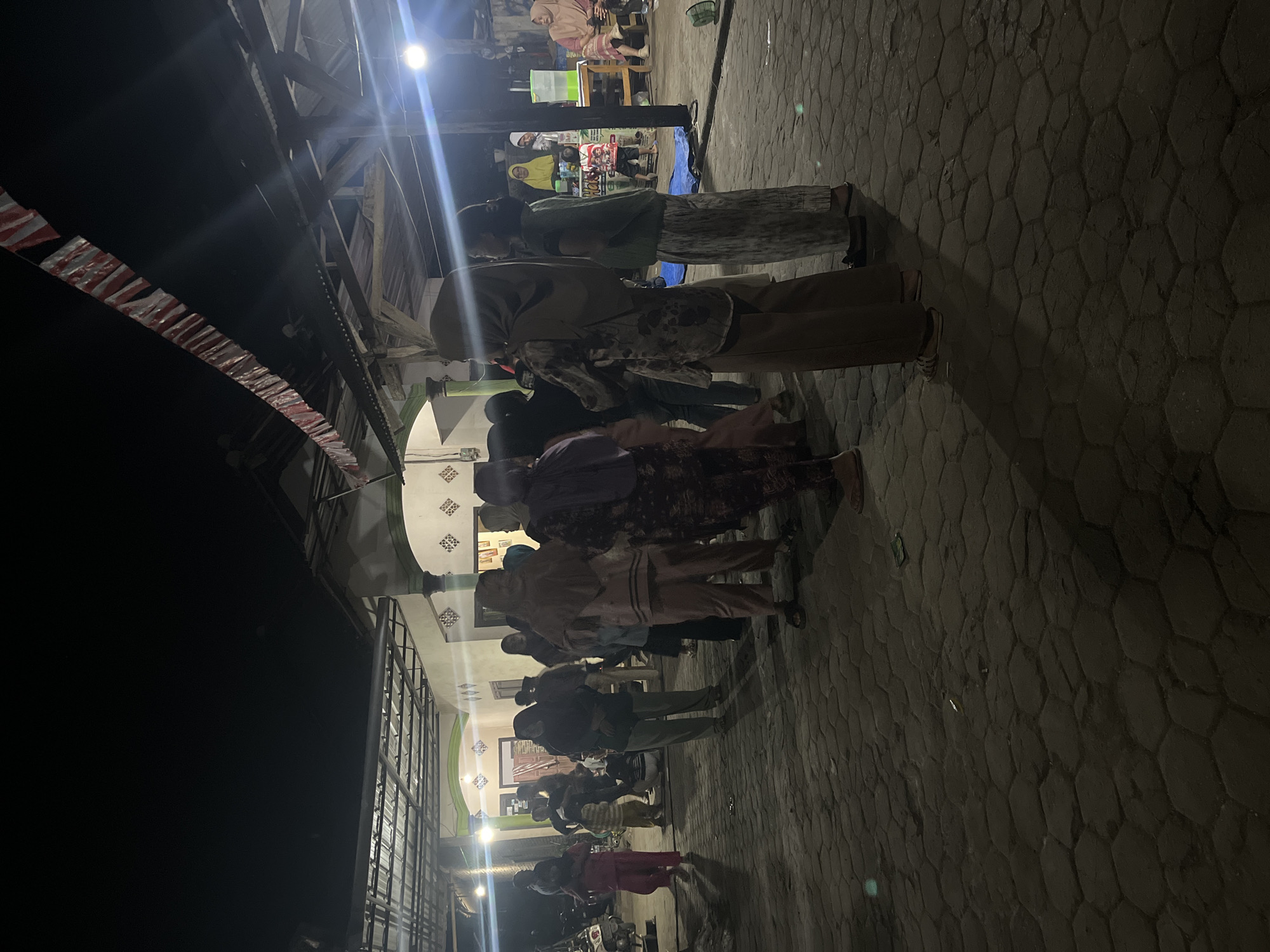 A group of people are gathered outdoors at night, in what appears to be a paved courtyard in Margodadi, Indonesia. The lighting is dim, with some artificial lights illuminating the scene and casting shadows. A building with a covered porch is visible in the background, adorned with red and white banners strung across the overhang. The ground is paved with interlocking hexagonal stones.
Several individuals are standing and walking, some are wearing headscarves and traditional clothing. A few are looking in different directions, while others appear to be in conversation. In the background, there are glimpses of what might be a food or drink stall with a large green dispenser. A motorcycle is parked to the left of the frame. The overall atmosphere suggests a community gathering or event.