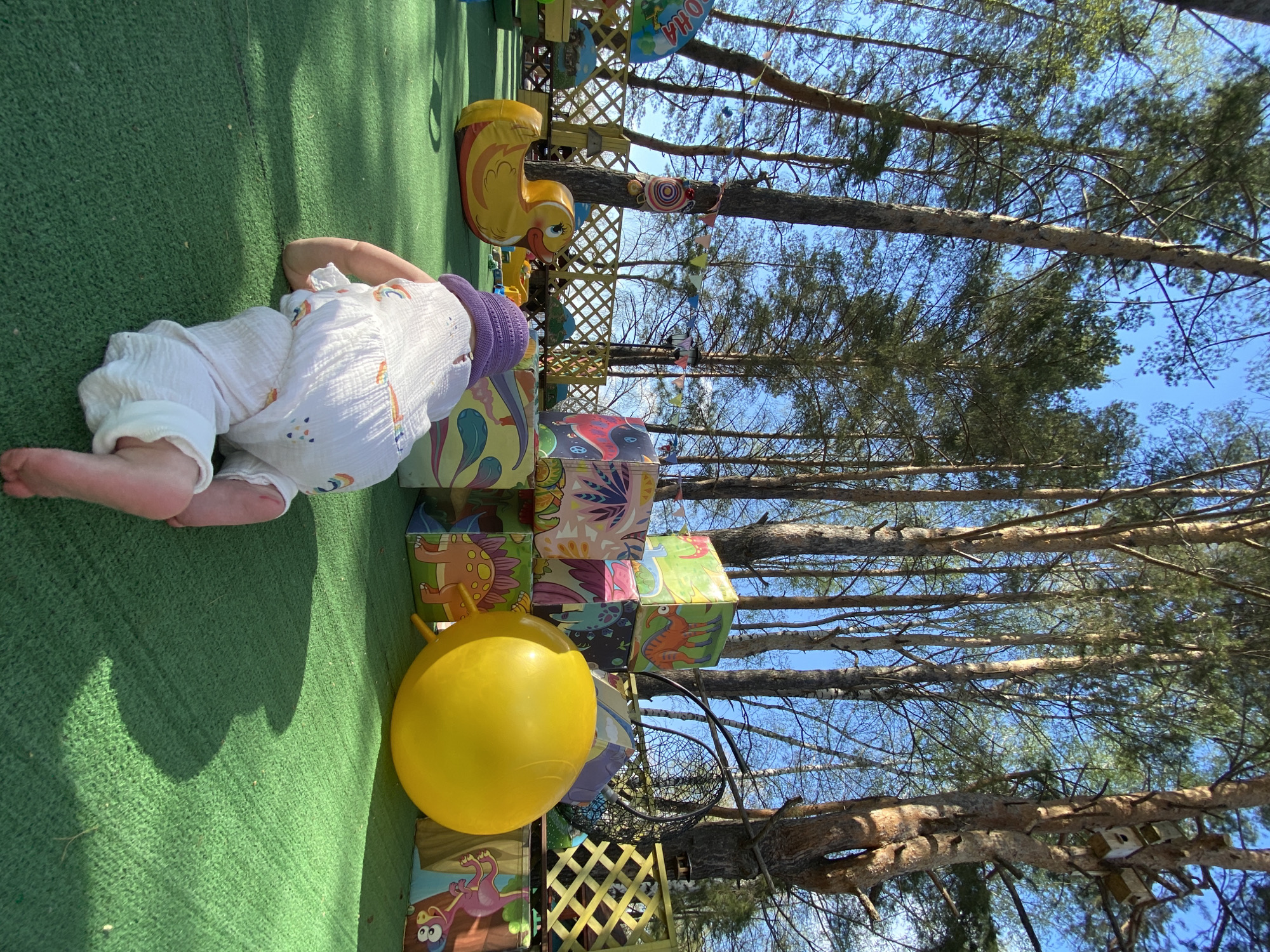 A baby wearing white clothing and a purple hat is lying face-down on green artificial turf. Beside the baby are stacks of colorful blocks with cartoon illustrations and a yellow inflatable ball. In the background are trees, a fence, and various decorations. The location is Chaikovsky, Russia.