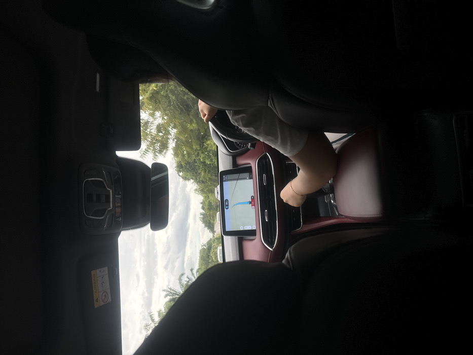 The image captures the interior of a vehicle, viewed from a rear passenger seat, in the city of Khian Sa, Thailand. A driver, identifiable by the back of their head and right arm, is visible in the front left seat. The driver is wearing a light-colored short-sleeved top; their left hand is on the steering wheel, and their right hand rests near the gear shifter on the center console. The vehicle's dashboard and central console are trimmed in a dark red material, accented with silver-colored details around the air vents. A large infotainment screen displays a navigation map with a blue route line, a vehicle icon, and numerical data including "40 km," "120 km/h," and a timestamp of "15:46." Above the dashboard, a rearview mirror and an overhead console are visible, along with an "AIRBAG" warning sticker affixed to the passenger-side sun visor. Through the windshield, an overcast sky with grey clouds is visible above a road flanked by dense green foliage, including palm trees on the left. Several white vehicles are discernible in the distance on the road ahead.