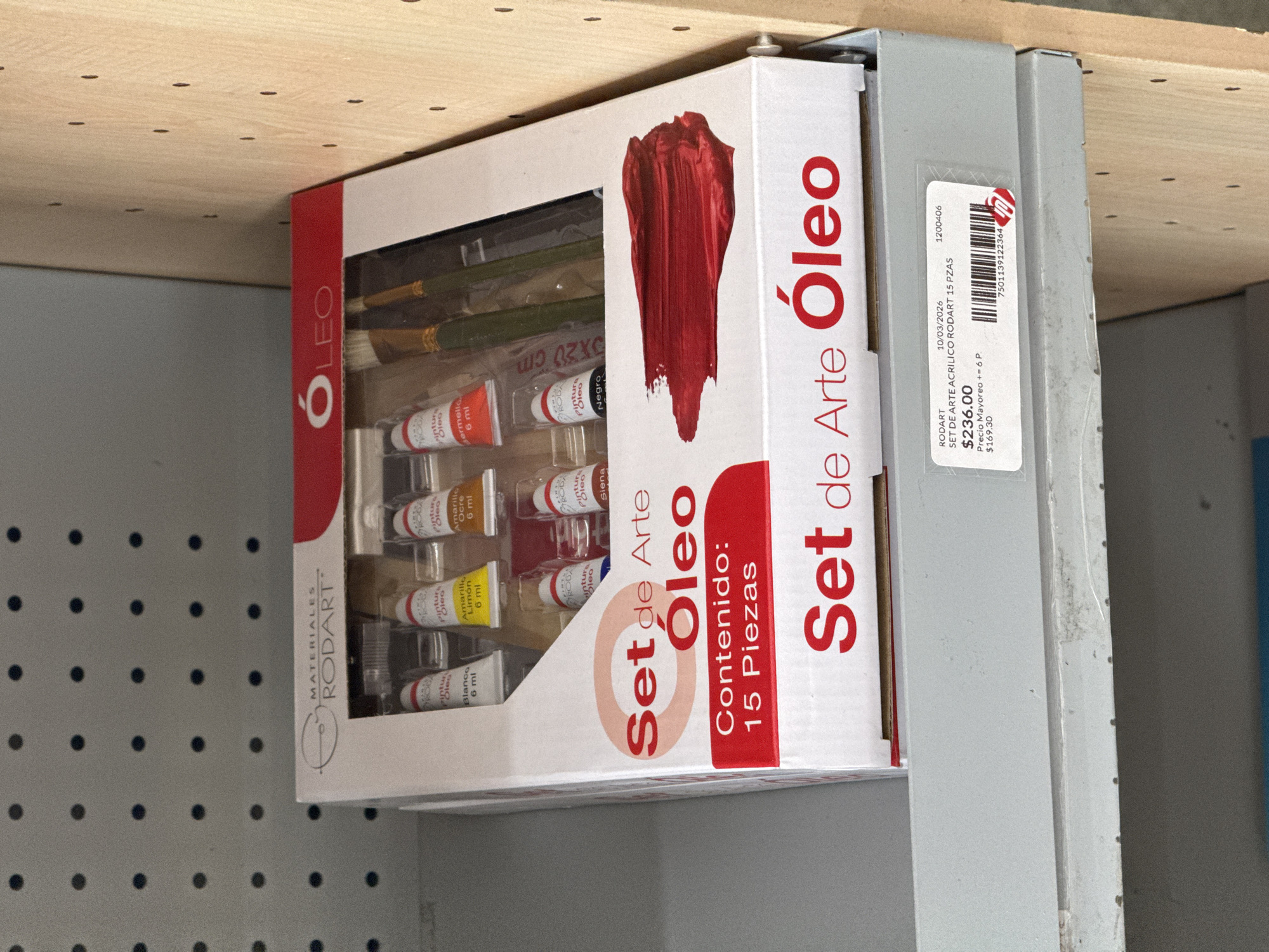 The image displays a "RODART" art set, specifically labeled "Set de Arte Óleo" (Oil Art Set), contained within a white cardboard box. The box is oriented vertically and sits within a grey metal display rack or shelf unit, likely in a retail environment in San Bernardino Tlaxcalancingo, Mexico.
The front of the box features a prominent red paint stroke design and a clear plastic window revealing the contents. Visible inside are multiple small tubes of "Pintura Óleo" (Oil Paint), each containing "6 ml", in colors such as "Blanco" (White), "Amarillo Limón" (Lemon Yellow), "Amarillo Ocre" (Yellow Ochre), "Siena", "Negro" (Black), and "Bermellón". Several paintbrushes are also part of the set. The box explicitly states "Contenido: 15 Piezas" (Contents: 15 Pieces).
A white price tag sticker is affixed to the metal shelf adjacent to the art set. The tag identifies the item as "SET DE ARTE ACRILICO RODART 15 PZAS", despite the box indicating "Óleo". The listed price is "$236.00", with a wholesale price ("Precio Mayoreo n= 6 P.") of "$169.30". The tag also shows a date "10/03/2026", an item number "1200406", and a barcode "7501139122364".
Above the metal display, a light-colored wooden panel with a grid of small dark holes is visible. The background consists of a plain grey wall. The scene is brightly lit, suggesting an indoor setting.