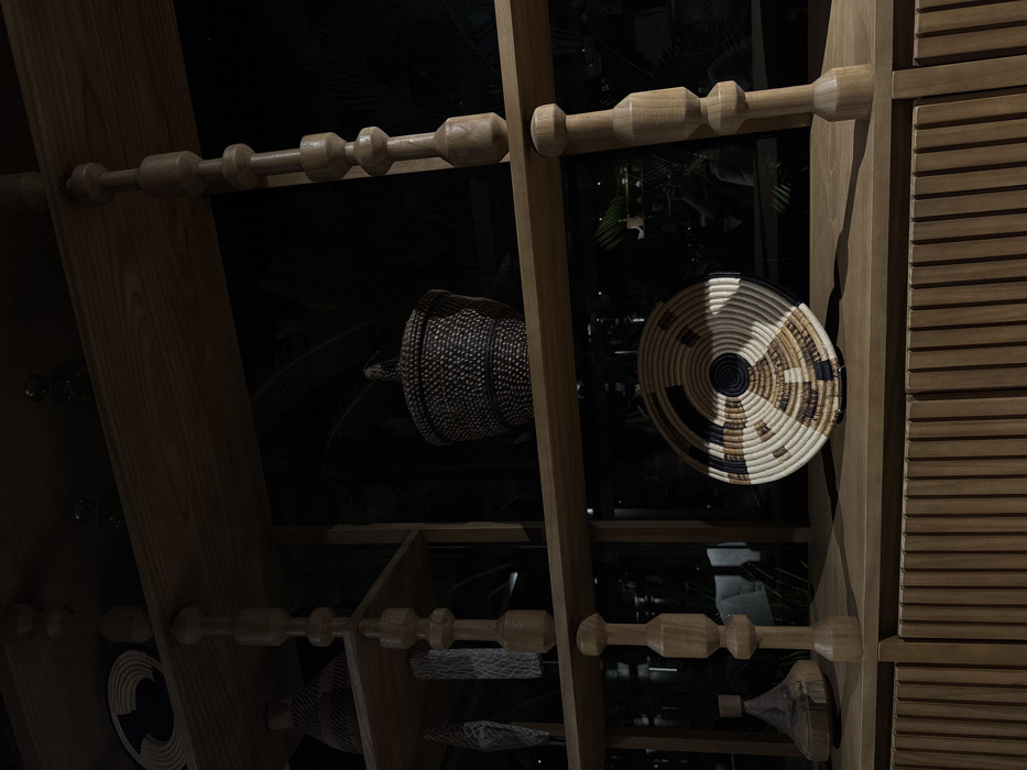 A slanted, open wooden shelving unit holds various woven baskets and vessels. A dark, globular basket with handles sits to the left of a flat, round woven piece with a black and tan concentric pattern. Horizontal wooden dowels with bulbous ends line the shelves. Vertical slats are visible on the right edge of the image. Dark foliage is visible through the open shelves.