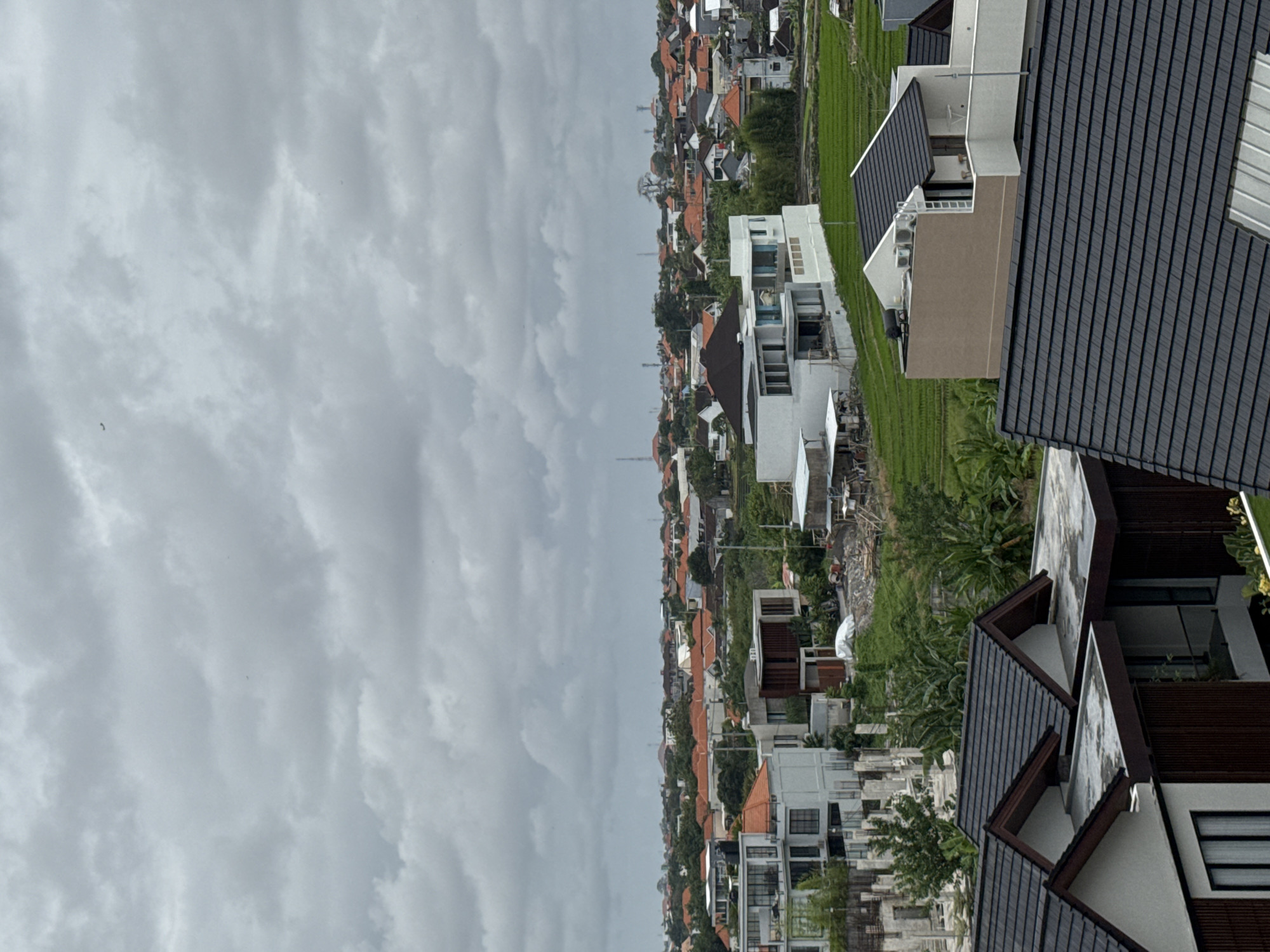 The image is a high-angle, medium shot of a residential area in North Kuta, Indonesia, under a cloudy sky. The foreground is dominated by the dark, tiled roofs of modern houses. In the midground, there is a collection of contemporary white houses with flat roofs, some featuring balconies and multiple stories, alongside older buildings with orange tiled roofs. Behind these houses, green rice paddies are visible, interspersed with trees and vegetation. Further in the distance, the landscape is a dense expanse of houses with orange tiled roofs, stretching towards the horizon. Several telecommunication towers are also present in the background. The overall scene depicts a suburban or semi-rural environment with a mix of new and older architectural styles.