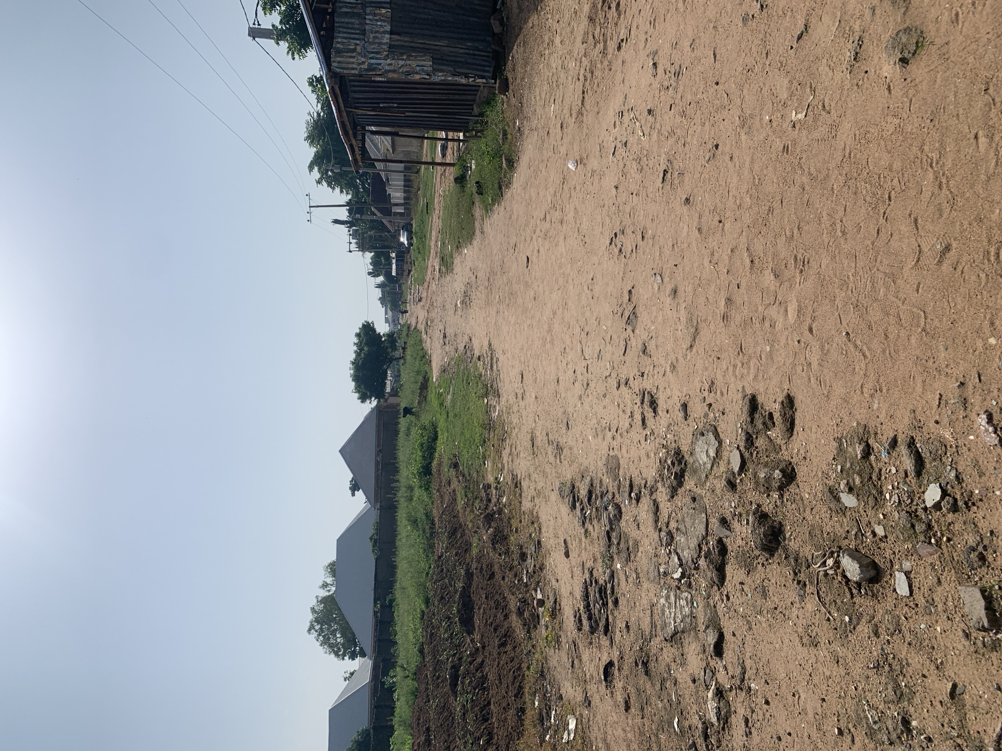 A sun-drenched, dirt road stretches into the distance, flanked by a mix of residences and corrugated metal structures. To the left, recently tilled earth adjoins a grassy area, beyond which several houses with grey, peaked roofs are visible. The road itself is uneven, marked by patches of exposed rocks and scattered debris, including plastic waste. On the right side of the road, a simple building with a corrugated metal roof and walls stands. Further down the road, utility poles with wires cross the sky, and several other houses and structures line the way. A single person is walking in the distance. The overall impression is of a dusty, rural or semi-urban environment under a clear, bright sky.