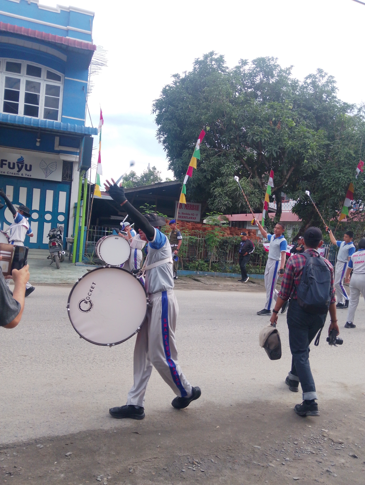 A street-level, eye-level medium shot captures a marching band parade in the city of Serbelawan, Indonesia. In the foreground, a young male, identified by the blue and white uniform and black hat, plays a large bass drum emblazoned with the word "ROCKET." He is facing slightly left, his right arm raised, holding a drumstick. To his right, another young male, also in uniform, stands with his arms raised, holding what appears to be a baton. Behind him, several other band members in similar attire are visible, some holding batons and drums. A person in a red and black plaid shirt with a backpack and camera walks away from the viewer on the right side of the frame. A police officer in uniform is visible in the background, standing near a brick wall. The parade is taking place on an asphalt road lined with buildings and trees. Colorful flags are strung across the street, some attached to poles. The building on the left features a blue facade with a white arched window and signage that reads "Fuyu Ice Cream & Tea." The sky is overcast.