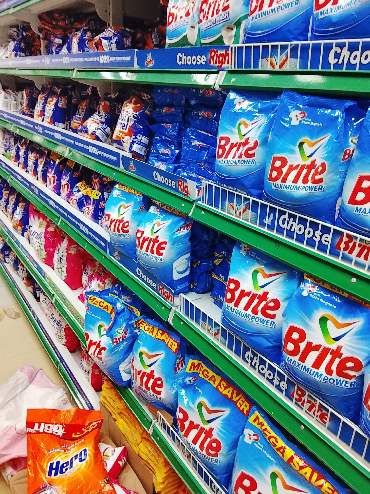 The image depicts a grocery store shelf in Gulgasht Colony, Pakistan, stocked with various laundry detergents. On the lowest shelf, an orange bag of "Hero" detergent is visible with an apparent price of 499 RS. Other detergents such as "Sunlight" are placed on the shelves above. Most prominent are the blue bags of "Brite" detergent, labeled "Maximum Power" and "Mega Saver." Blue shelf dividers with "Choose Right" are visible. There is a white bag on the floor next to the "Hero" detergent. The shelves are green and white.