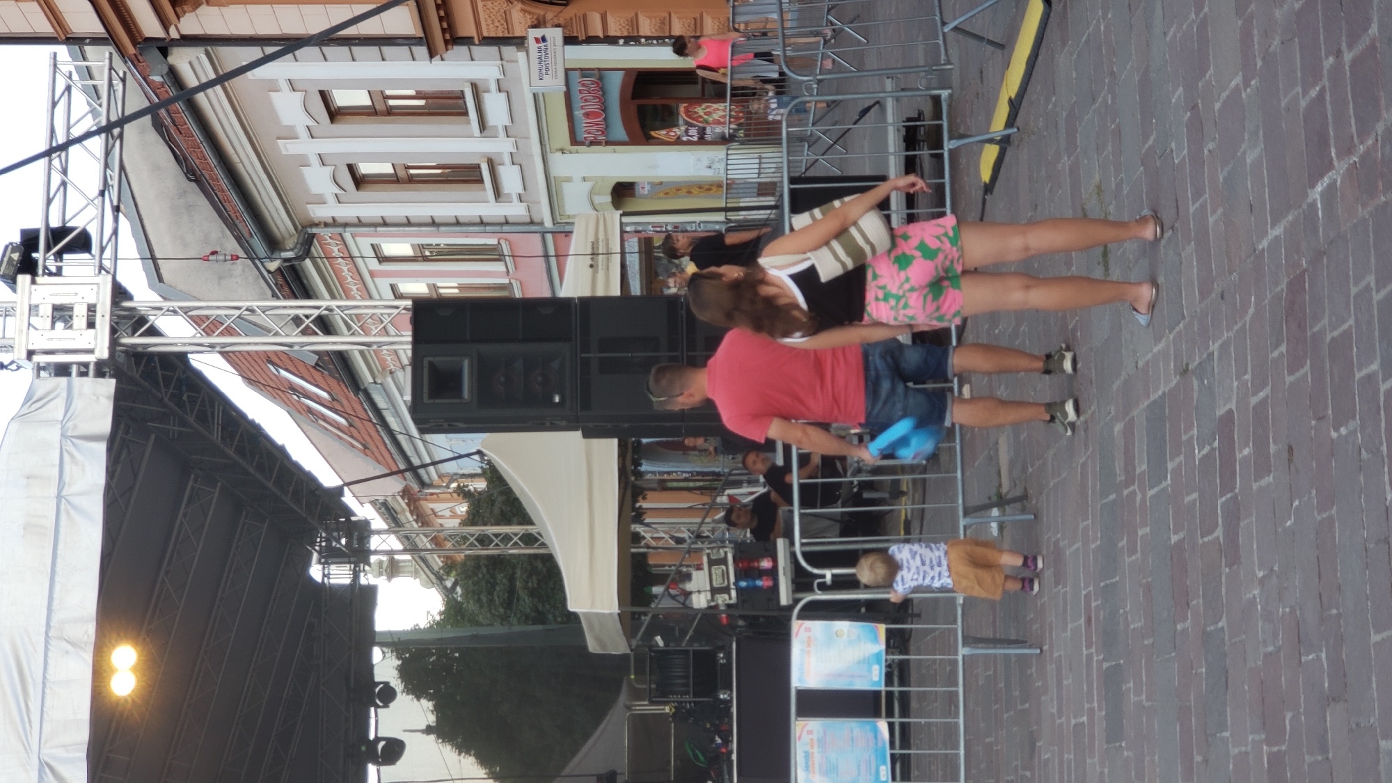 The image depicts an outdoor event with a stage and sound equipment. In the foreground, a young child wearing a patterned shirt and orange shorts stands facing away from the camera. To the right, a man in a red polo shirt and blue denim shorts holds a blue inflatable object. Next to him, a woman in a black and white top and pink floral shorts with a striped bag over her shoulder is also facing away. They are standing behind a metal barrier. In the background, there are buildings with traditional architecture, a white tent, and more people. The ground is paved with brick. The scene appears to be a public square or street.