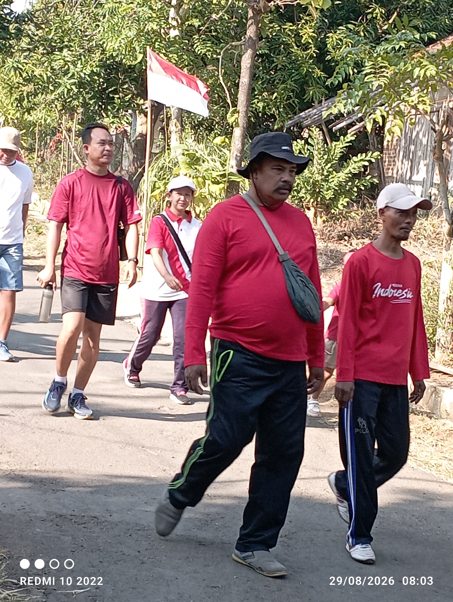 The image depicts a group of people walking on a paved road lined with vegetation and buildings. The scene is likely in Dukuhseti, Indonesia, as indicated by the timestamp. Several individuals are visible, most wearing red shirts, suggesting a group activity or event. One person in the foreground wears a red long-sleeved shirt with "Indonesia" printed on it, a black hat, and a green bag slung across their chest. To their right, another person in a red long-sleeved shirt and a white cap is walking. In the background, an Indonesian flag flies. The overall atmosphere appears casual, possibly a morning walk or a community event. The environment suggests a rural or semi-urban setting.