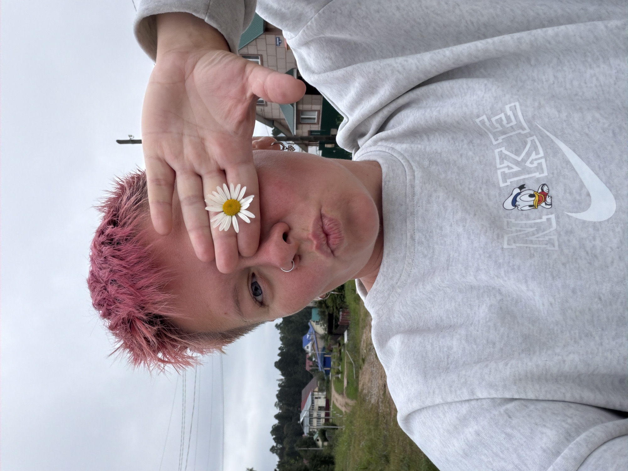 A medium shot shows a person with short, pink-dyed hair, holding their left hand up to cover their left eye and face. A daisy is placed on the person's outstretched fingers, with its petals extending over the eye. The person is wearing a light gray Nike sweatshirt with a graphic of Donald Duck. They are looking at the camera with their mouth pursed, and a nose ring is visible. The background is a slightly blurry outdoor scene, depicting a rural or suburban area with several houses, trees, and a dirt road. The sky is overcast and gray. The image is taken in the city of Chaikovsky, Russia.