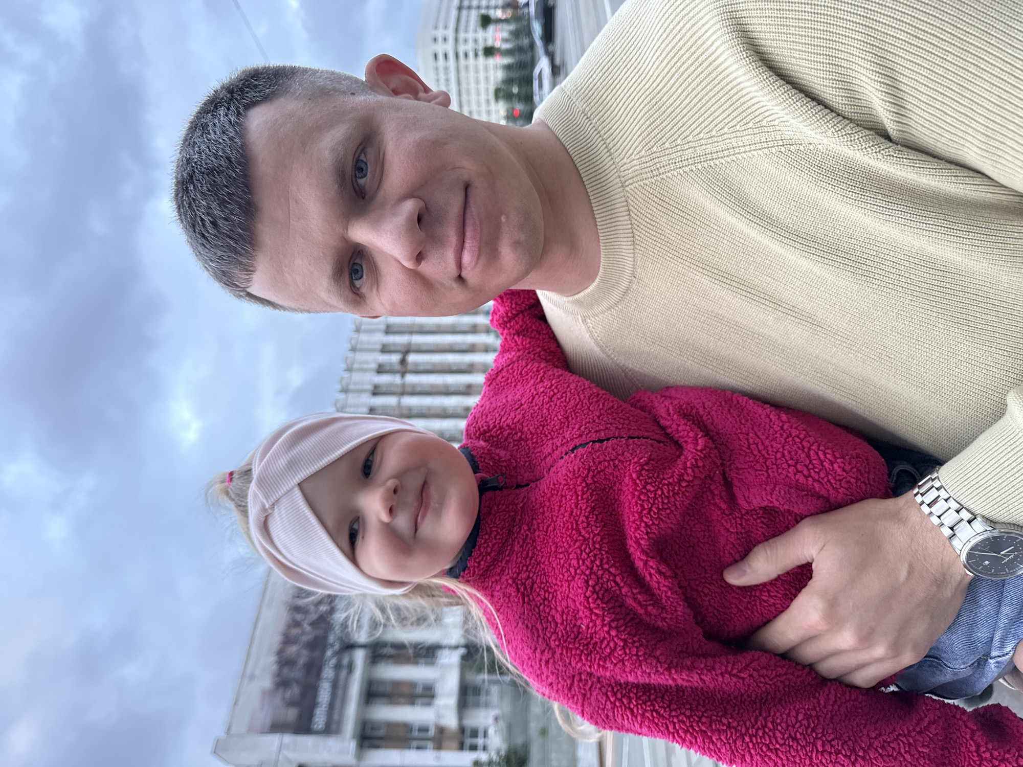 A man is holding a child in his arms. The man has short gray hair and is wearing a light-colored sweater. The child has blonde hair with a pink headband and is wearing a pink fuzzy jacket. The man is wearing a watch on his left wrist. They are standing in an urban setting. In the background are buildings and trees. The building has a sign visible with the first word “South". They are located in Perm, Russia. The image appears to be taken from a low angle. The sky is overcast.