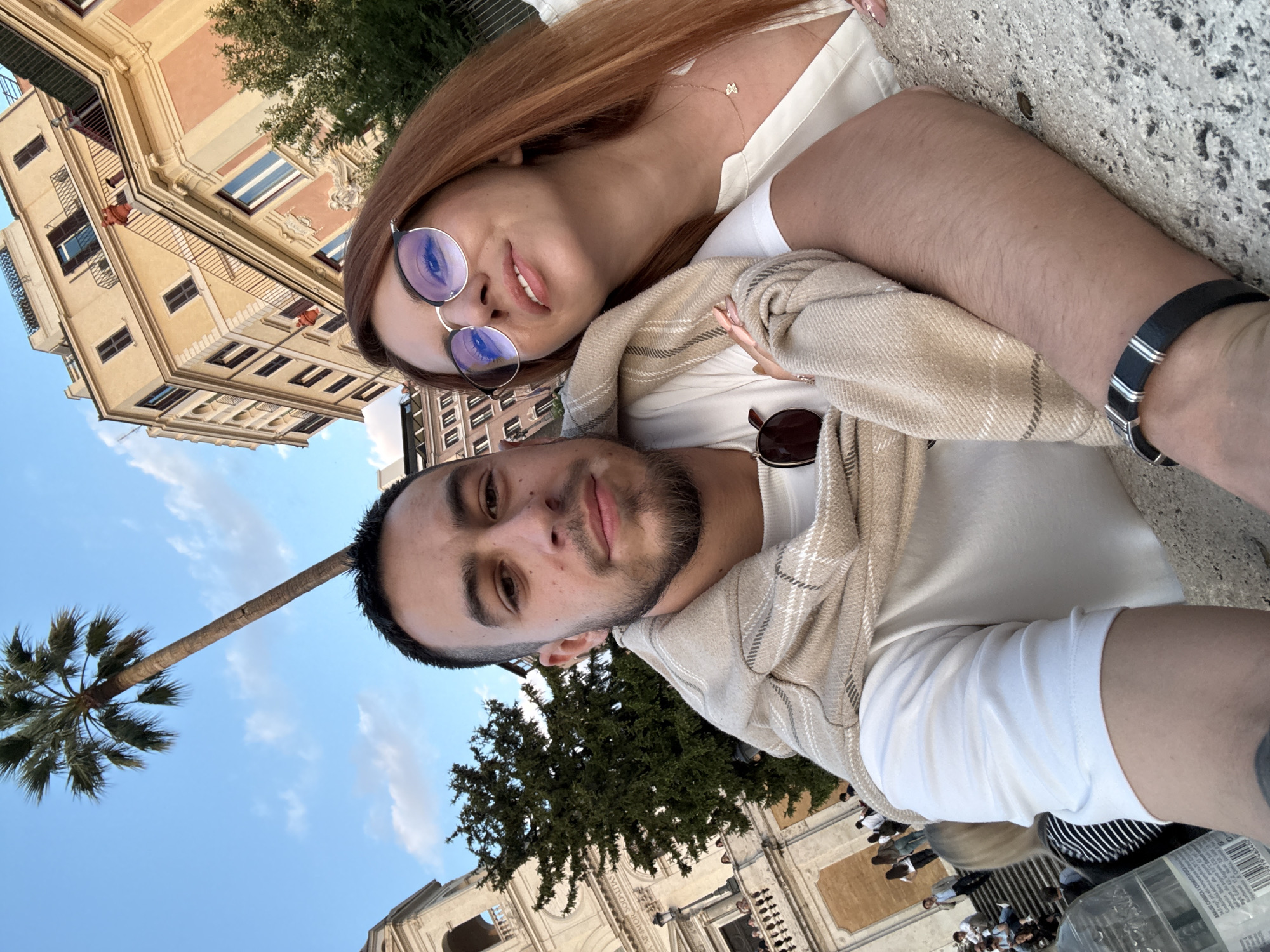 A medium shot, taken from a low angle, shows two individuals in an embrace. The female has shoulder-length red hair, wears round glasses, and is dressed in a white top. The male, sporting a mustache and short dark hair, is also in a white shirt with a beige shawl draped over his shoulders. He wears a black and silver bracelet on his right wrist.
In the background, there's a clear blue sky with a few clouds, a tall palm tree, a grand European-style building with several stories and numerous windows, and other trees. A crowd of people can be seen further in the background.
Based on architectural style and other visual cues, it is likely that the photo was taken in Rome, Italy, near the Piazza Navona.