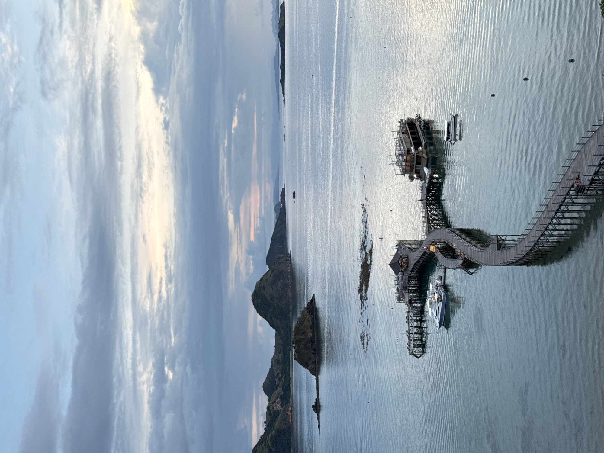 The image depicts a seascape in Labuan Bajo, Indonesia, under a cloudy sky with subtle hints of sunset. The water is calm with slight ripples. A long, winding wooden pier extends from the right side of the image into the water, leading to a structure with steps down to the water level. A large white boat is docked at this structure, while further in the distance, another more primitive-looking wooden platform or small dock with boats is visible. Several small islands with vegetation can be seen dotting the horizon, and the background suggests mountainous terrain. No people are clearly visible, but the pier appears to be designed for pedestrian traffic.