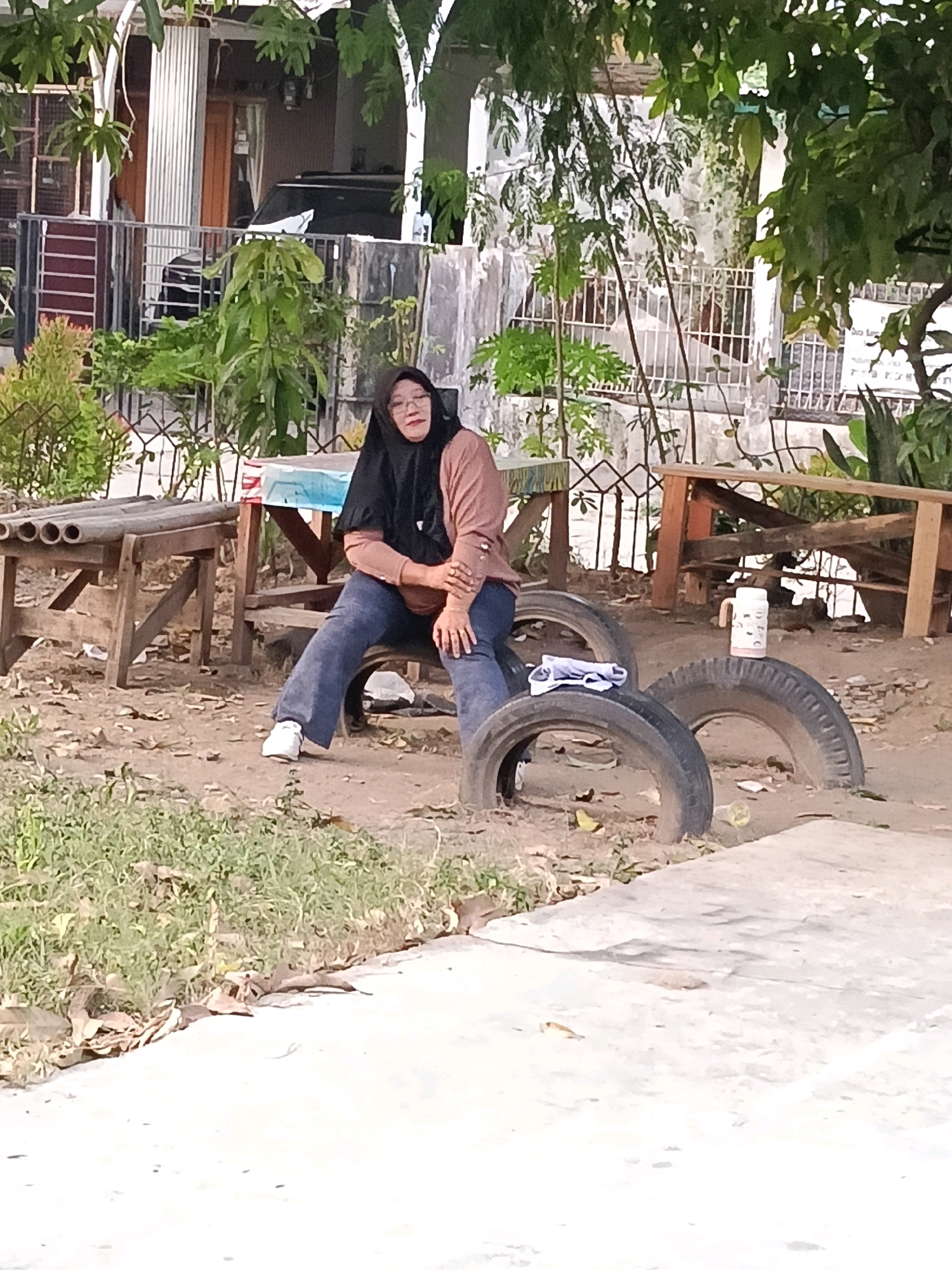 A woman wearing a black hijab and a brown long-sleeved shirt is sitting on stacked tires in what appears to be an outdoor area in Jatimulya, Indonesia. She is also wearing blue pants and white sneakers. In the background, there is a fence, foliage, and a building with a car parked in front of it. There are also some wooden benches and more tires scattered around the ground. A white insulated bottle with a pattern of cartoon animals is placed on one of the tires. The ground is dirt and covered with some dry leaves.