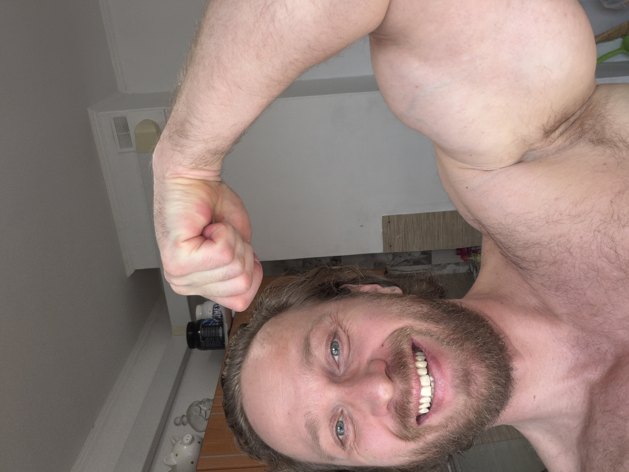 An indoor photograph features a smiling adult male, appearing Caucasian, with light skin, blue eyes, and light brown hair and beard. He is shirtless, exposing his chest and shoulders, and actively flexing his left bicep (viewer's right arm), with his hand fisted.
The immediate background displays a light-colored wall and a white ceiling with decorative crown molding. To the subject's left, a wooden or laminate cabinet is partially visible. Above the subject's head, on a shelf, are two white ceramic pig figurines with gold spots, a dark container, and a white container clearly marked "WHEY." A white rectangular ventilation grille is visible on the upper wall or ceiling, situated above a small, light-colored, possibly beige, wall fixture. The environment suggests a private residence. The specific location within Saint Petersburg, Russia, cannot be determined from the visual information provided.