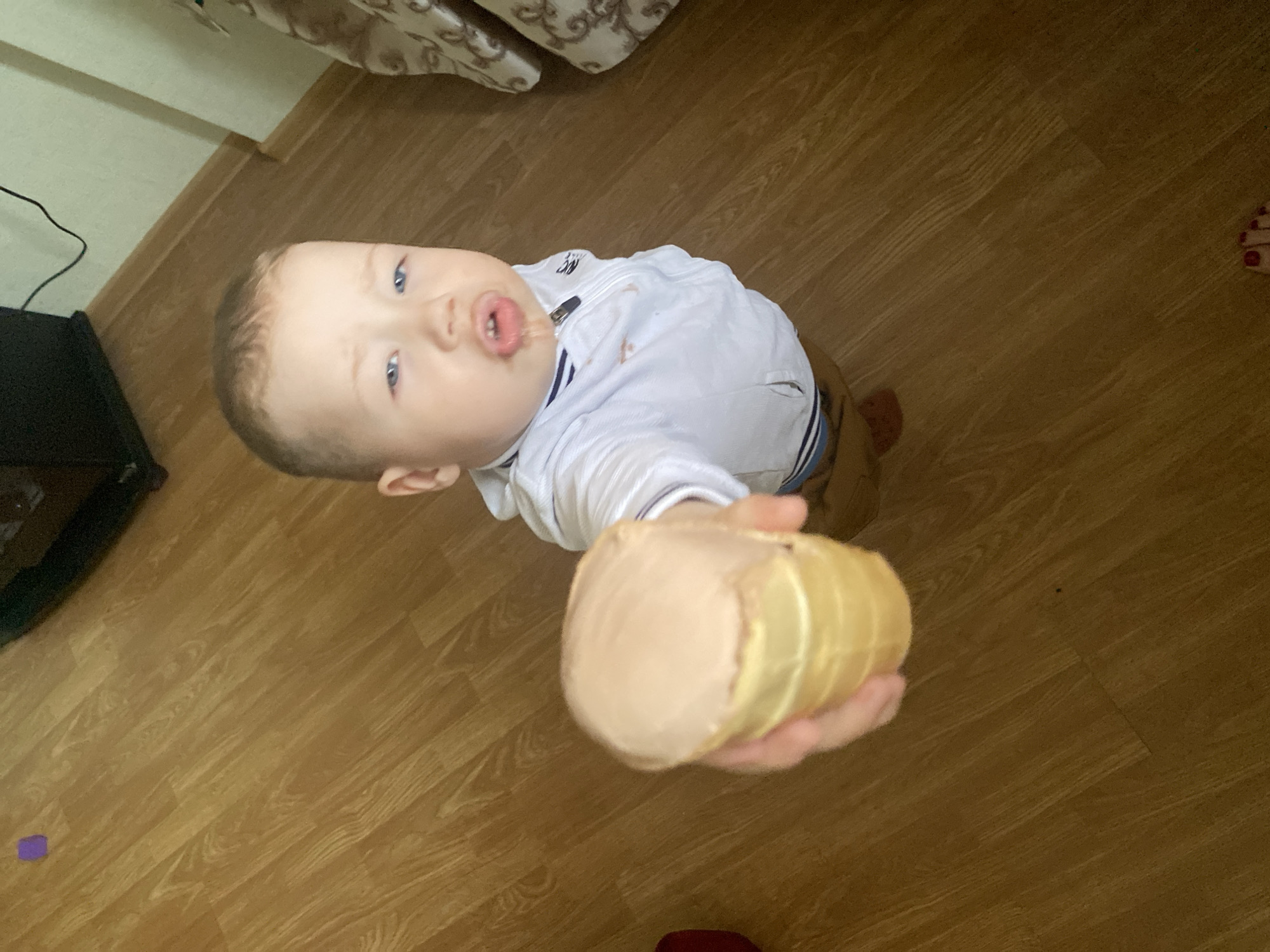 A top-down, medium-shot shows a light-skinned baby holding out an ice cream cone. The baby has blue eyes and is wearing a light-colored collared shirt with dark stripes and khaki pants. The ice cream cone is cylindrical with light brown ice cream on top and a light yellow cone. The floor is a light brown wood laminate. Parts of a bed with beige patterned bedding are in the upper left corner of the image. A woman's foot is in the upper right corner. A television and its power cord are in the lower left. The location is unknown and not identifiable as Sochi, Russia.