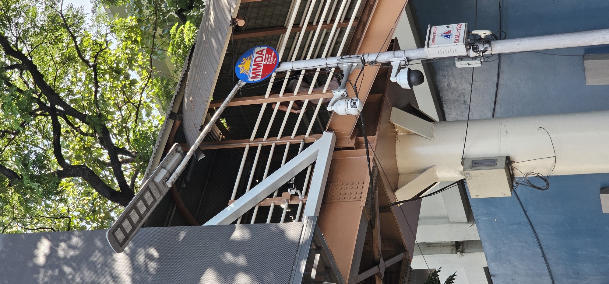 The image shows a segment of an elevated pedestrian walkway in Quezon City, Philippines, on a sunny day. The walkway is supported by thick, painted steel beams. A solar-powered street lamp is affixed to the upper structure of the walkway. Below the walkway, a pole holds two security cameras, one higher than the other. A circular sign with the MMDA logo and text in Filipino, indicating an emergency hotline, is mounted on the same pole. Electrical boxes and wiring are visible on and around the support structures. Lush green trees with dense foliage are present in the background, casting shadows on the walkway. The pedestrian walkway itself is constructed with white railings and a corrugated metal roof. The lower portion of the scene includes a blue-painted wall.
