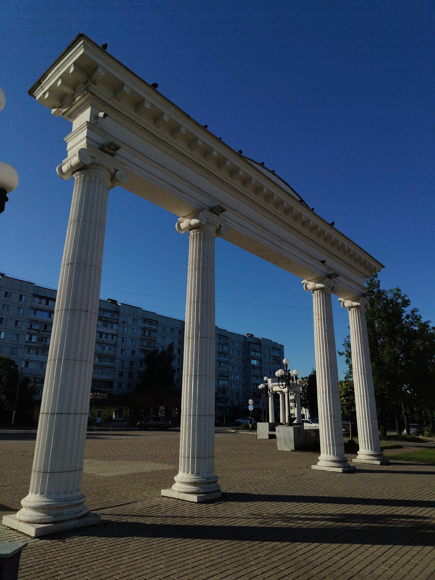 The image captures a low-angle view of a white, colonnade-style architectural structure composed of five fluted columns supporting a horizontal entablature. The structure is situated in a public square, identified by brick-paved ground and an urban backdrop. The sky is a clear, deep blue. Buildings are visible in the background, revealing an urban setting. Shadows are cast across the brick pavement, indicating the presence of strong sunlight. Trees with green foliage are also seen, suggesting a park or green area adjacent to the square. The photograph lacks any signs of human presence or activity. The location is in the city of Unknown, Unknown.