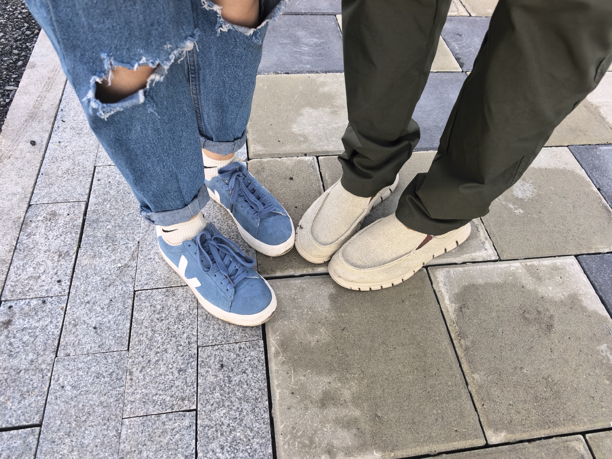 Two individuals are positioned on a paved outdoor surface, with the view captured from above, focusing on their lower legs and footwear.
The person on the left wears light blue denim jeans, cuffed above the ankles, with a prominent tear above the left knee exposing skin. They wear white crew socks featuring a black Nike swoosh logo on the outer ankle. Their footwear consists of blue suede low-top sneakers with white laces and a white "V" logo on the lateral side, set on white rubber soles.
The person on the right wears dark olive green full-length trousers, which are slightly bunched around the ankles. They wear light beige or off-white textured slip-on shoes with white, ridged soles. A subtle reddish-brown stripe is visible on the inner side of the right shoe's sole.
The ground surface is a patchwork of pavers. On the left, the pavers are rectangular and light grey, exhibiting a granular texture consistent with granite. Towards the center and right, the pavers are larger, predominantly light grey to off-white concrete-like slabs, showing minor discoloration and moisture patches. The two individuals are standing with their feet relatively close, suggesting they are either stationary or engaged in an interaction. The lighting is bright and even, indicative of daylight.