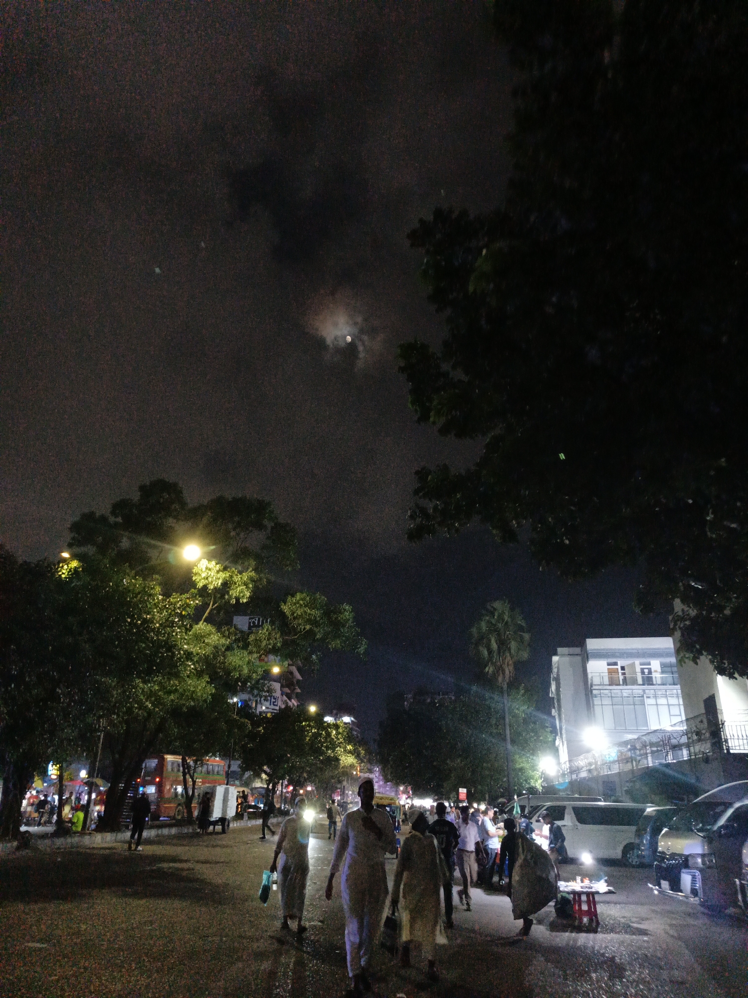 The image depicts a street scene at night in Dhaka, Bangladesh. A paved road runs diagonally from the bottom left towards the upper right. Several people are walking on the road, some of whom are carrying bags or pushing carts. A brightly lit street lamp illuminates a large tree on the left side of the road. Further down the street, there are more trees, buildings with illuminated windows, and various vehicles, including a red bus and vans. The sky is dark with clouds and a faint moon is visible. The overall atmosphere is of a bustling urban environment at night.
