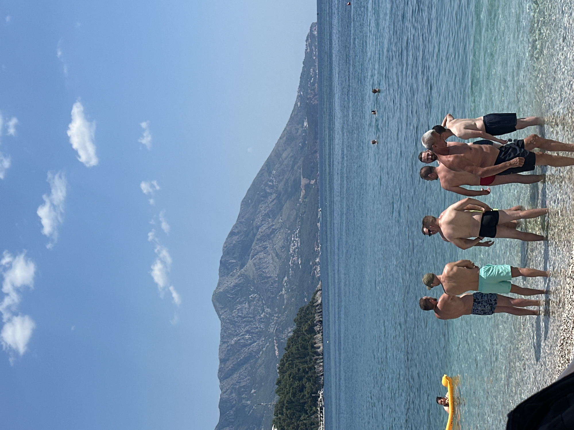 This media file shows a group of people on a beach in Budva, Montenegro. The scene is set by a clear blue sky with a few scattered clouds and a large, rocky mountain in the background. The water is clear and shallow, with a pebble beach. Several men are in the water, some standing and talking, while others are partially submerged. One man is on a yellow inflatable raft. The overall impression is a sunny day at the beach, with a relaxed and casual atmosphere. The time of day appears to be midday, given the bright sunlight and the shadows. The location is suggested by the distinctive mountain landscape typical of the Budva Riviera. There is no visible text in the image.