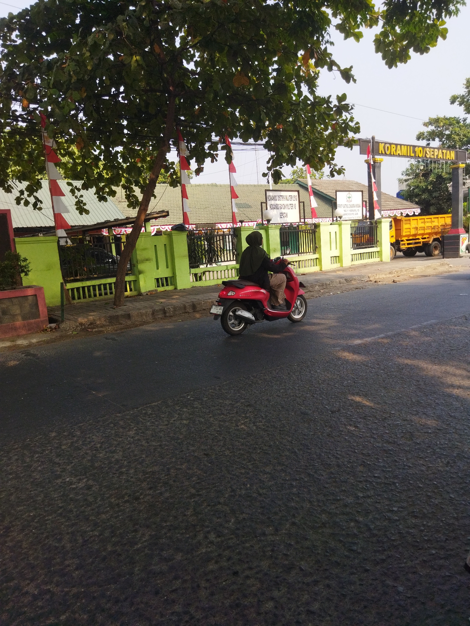 A person rides a red scooter in Sepatan, Indonesia, on a sunny day. The scooter is traveling on a paved road with the person wearing a dark green headscarf and patterned pants. In the background, a green building with a white roof is visible, adorned with red and white Indonesian flags. A sign above an archway reads "KORAMIL 10/SEPATAN". To the right of the building, a yellow truck is parked. The scene is captured from a low angle, with a large tree with green leaves dominating the upper left portion of the frame. The sky is a clear blue.