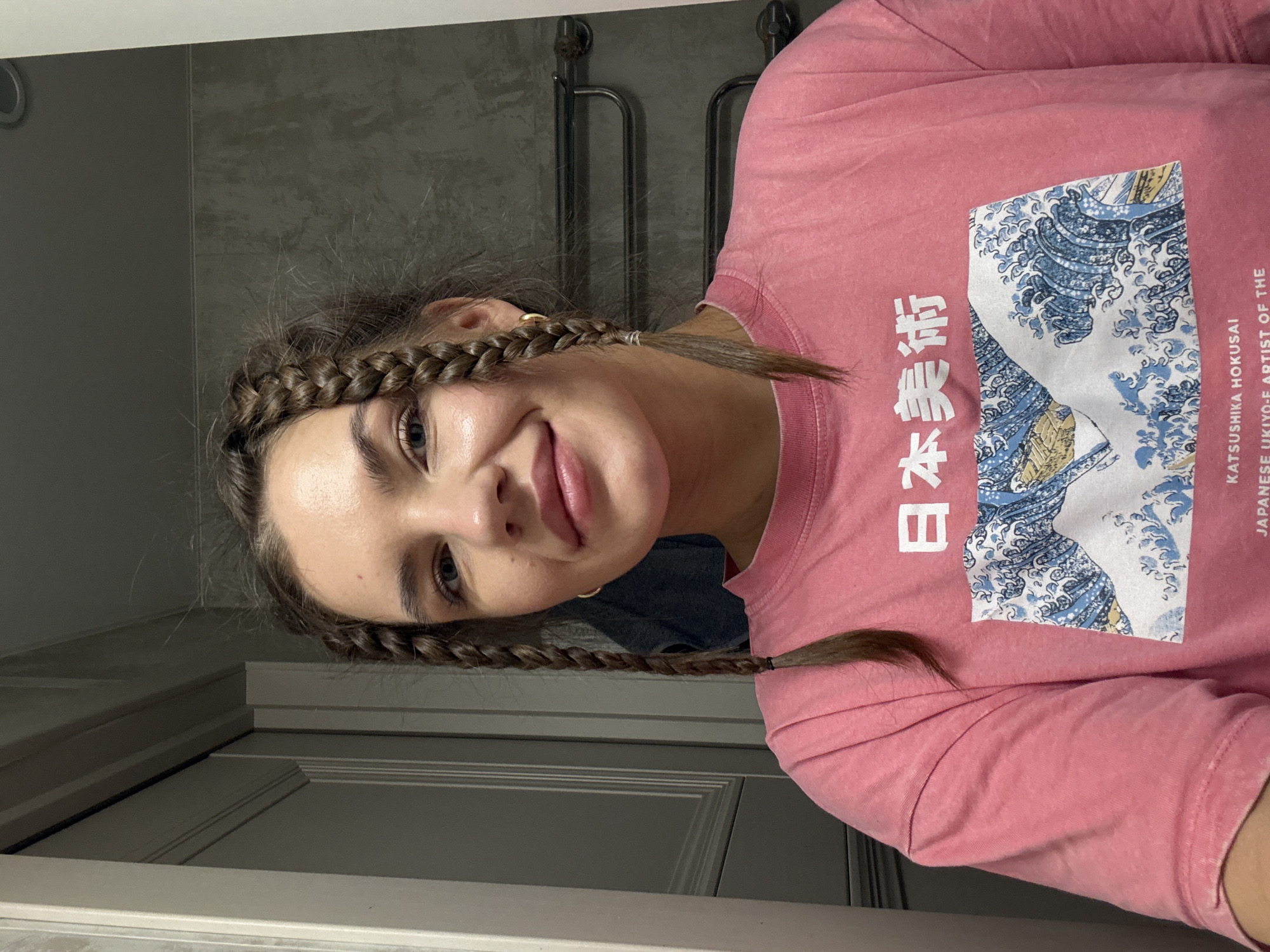 A young woman is posing for a selfie in what appears to be a bathroom. She has brown hair styled in two braids. She is wearing a pink t-shirt with a graphic of "The Great Wave off Kanagawa" by Hokusai. The background shows a bathroom door and gray tiled walls with a metal towel rack. There is no evidence to suggest this image was taken in Moscow, Russia.